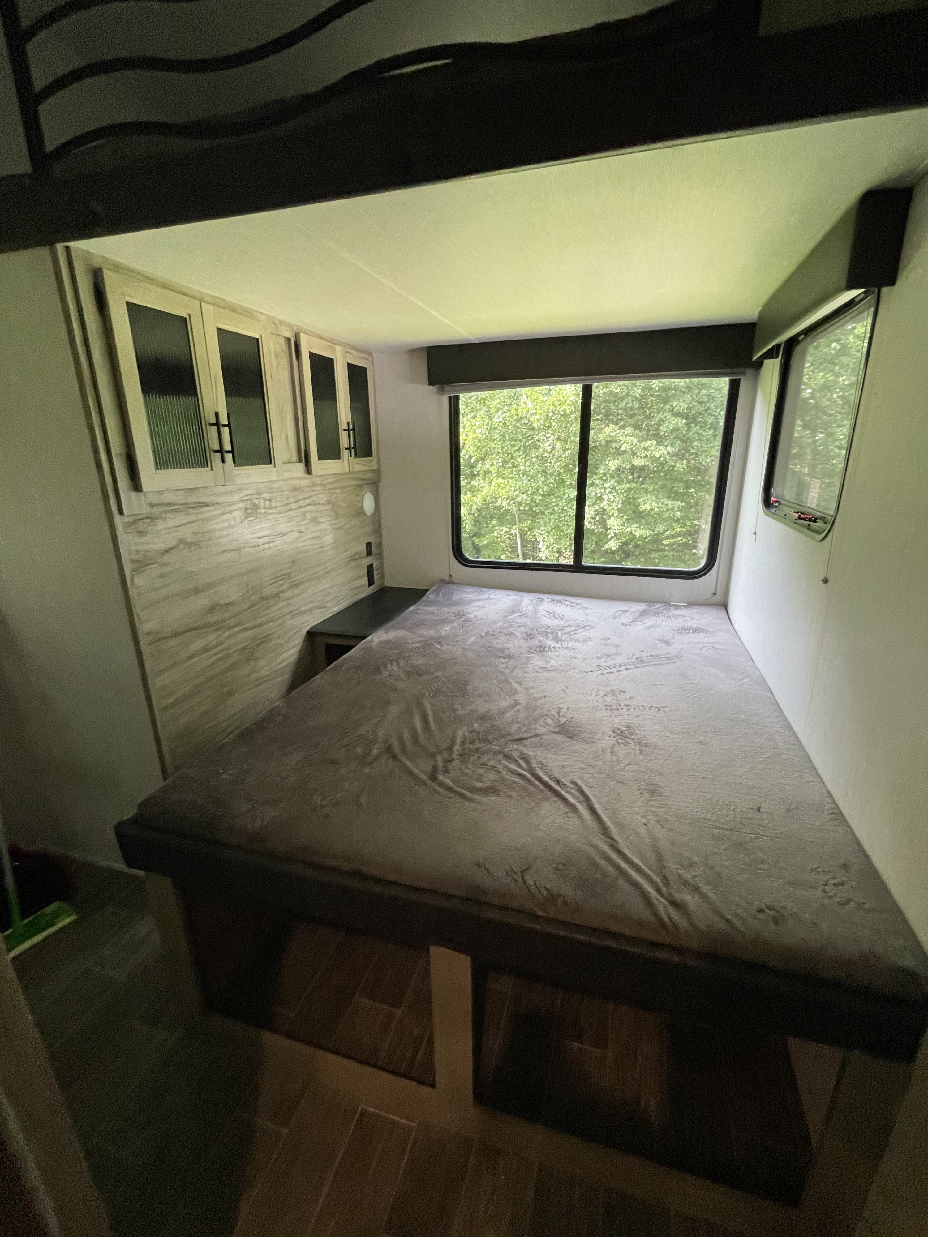 Inside an RV rental bedroom: a large bed with grey blanket, window with trees outside, and overhead cabinets with wood paneling.. Forest River Cherokee Arctic Wolf 2022