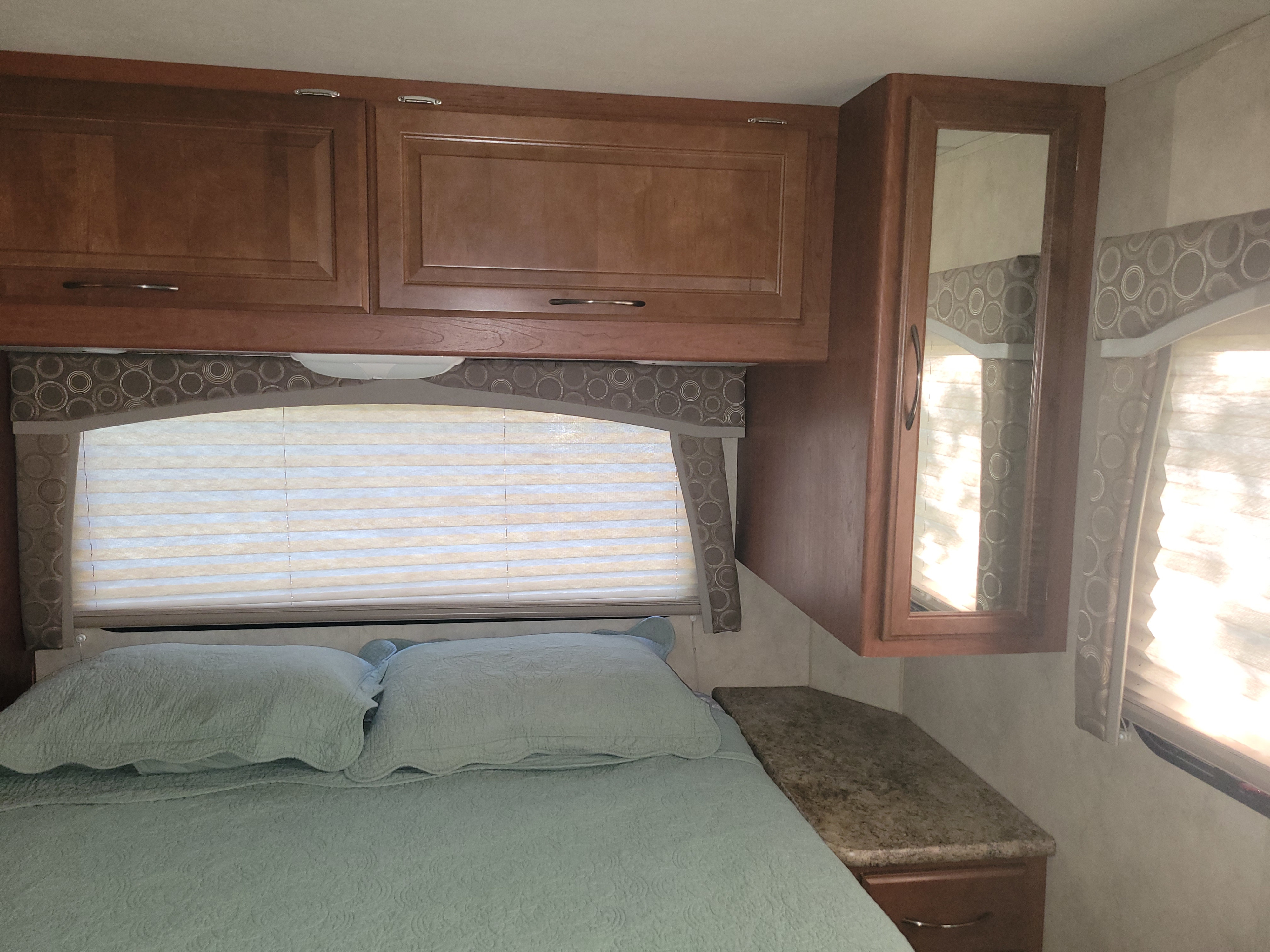 RV rental bedroom with wood overhead cabinets, a light blue bed, window blinds, and a mirrored cabinet beside a nightstand.. Thor Chateau 2012