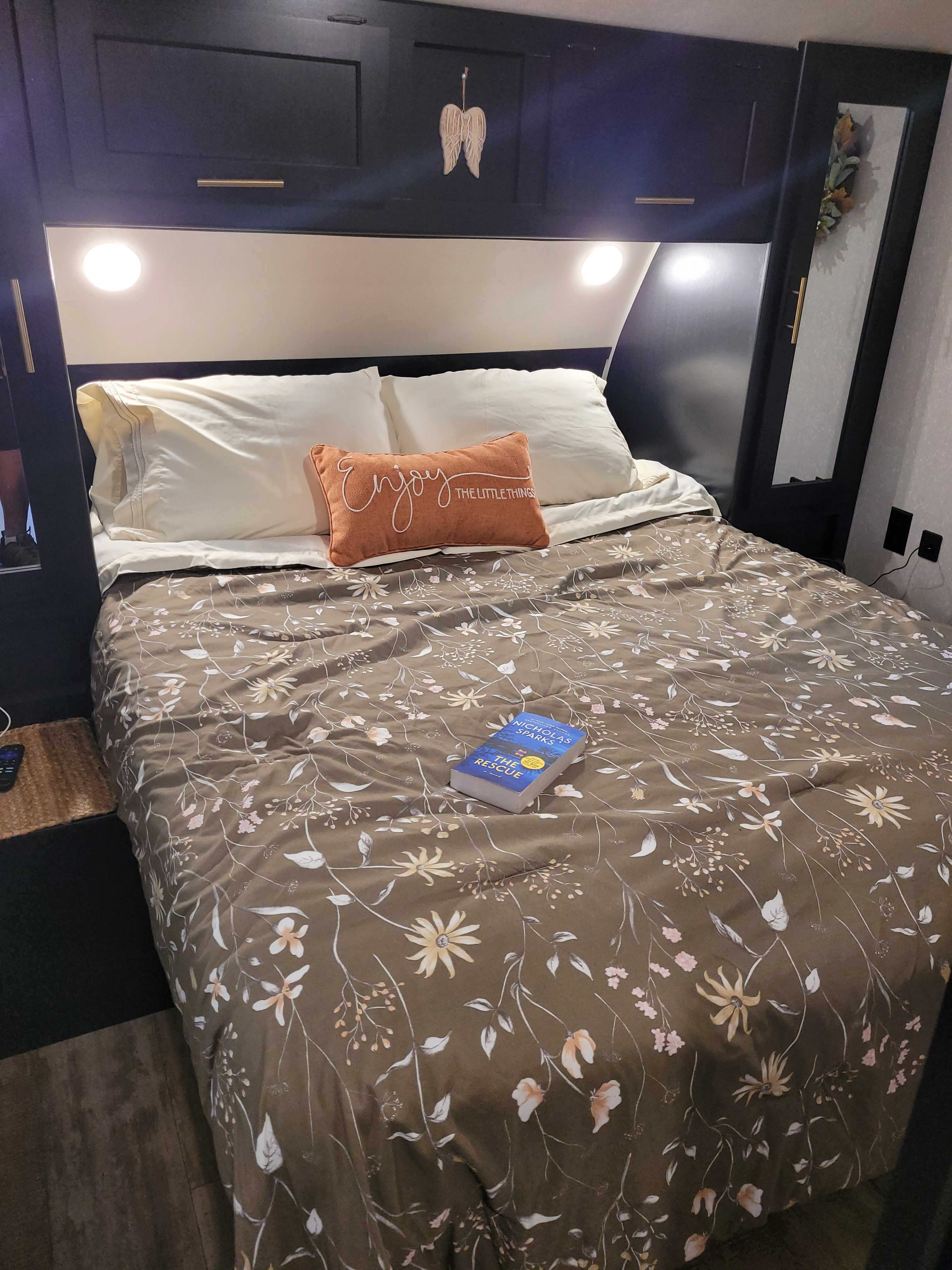 Relax in this RV rental bedroom: a cozy bed with floral comforter, pillows, and a book. Overhead cabinets & lighting.. Forest River Cherokee 2021