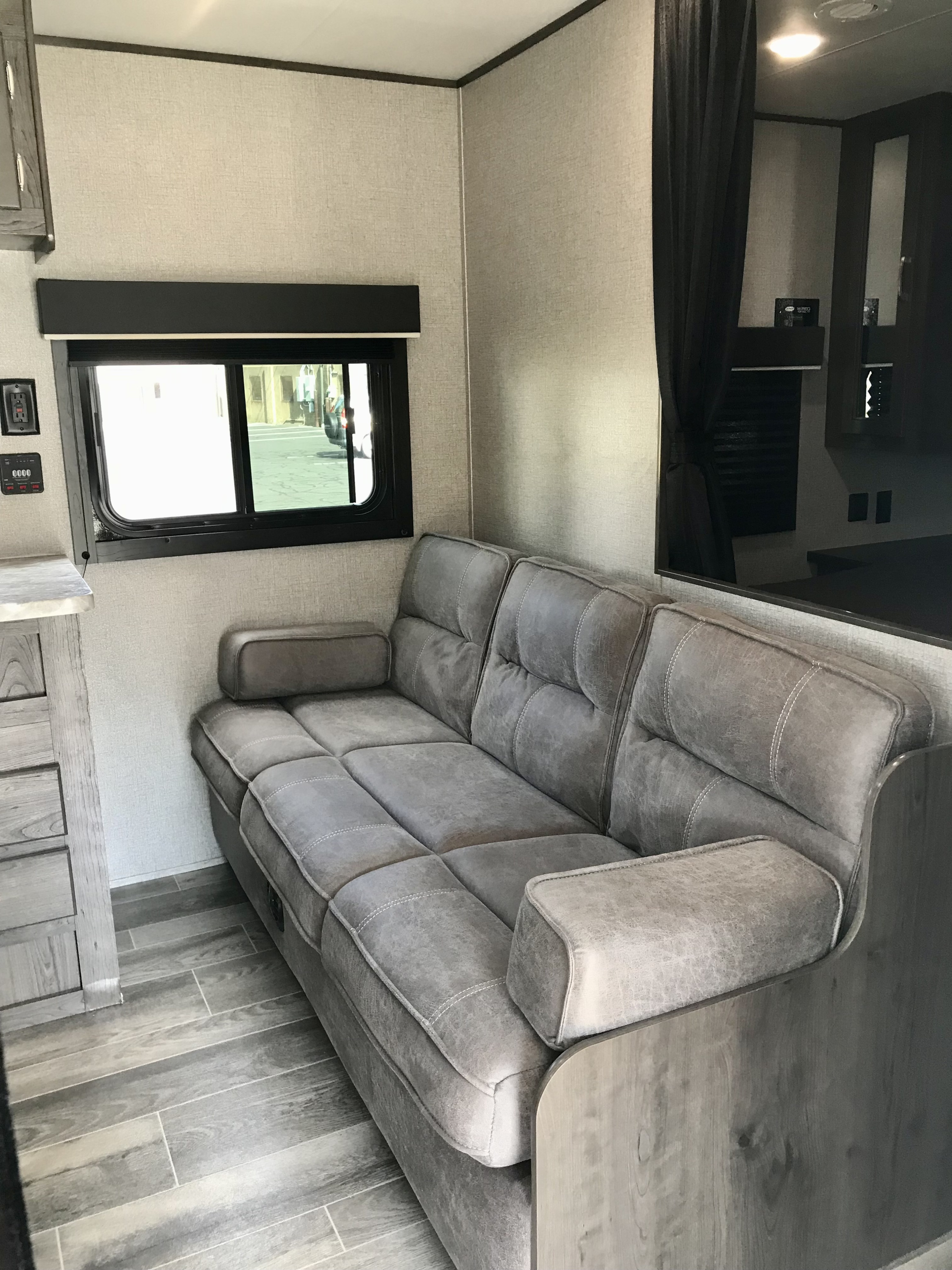 Inside a modern RV rental: A grey sectional sofa, wood-look floor, and window create a cozy living space.. Jayco Jay Flight SLX 2021