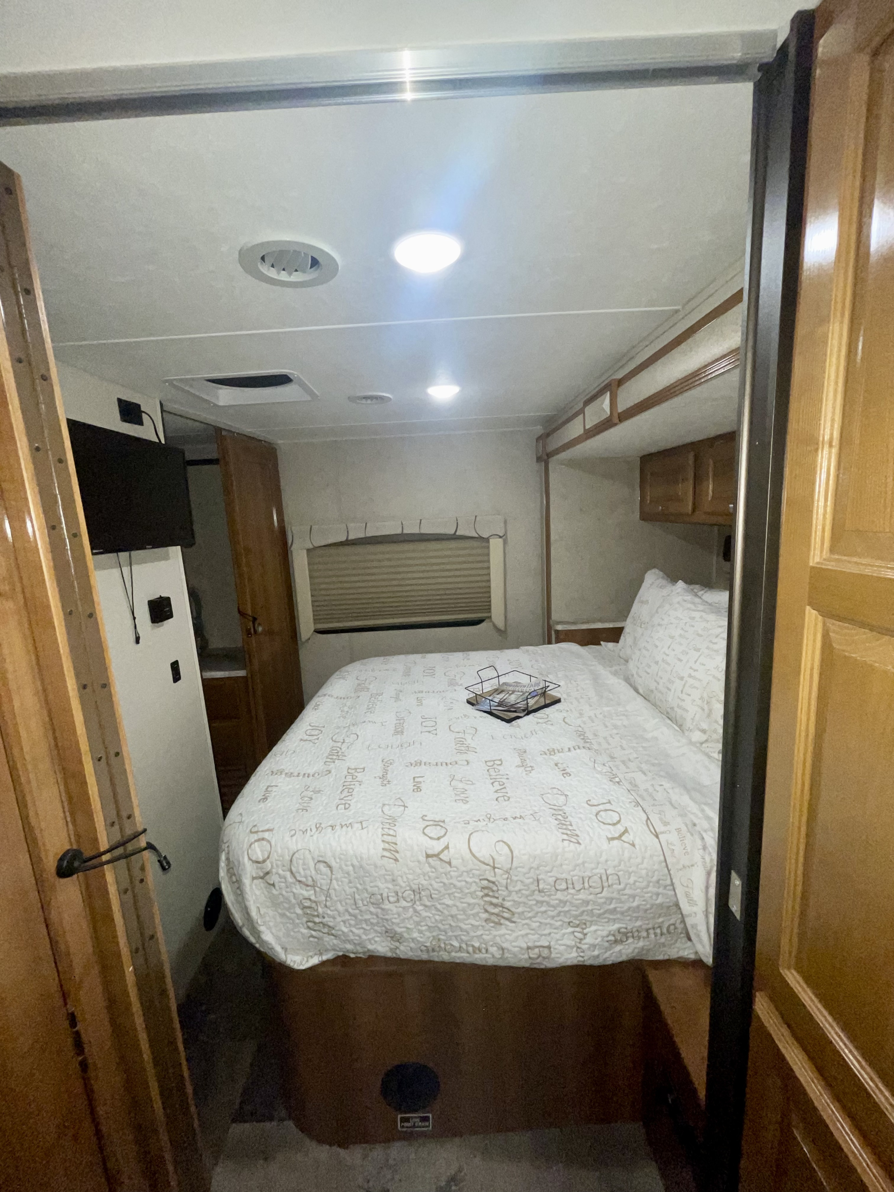 Cozy RV rental bedroom interior with a comfortable bed, overhead lighting, wood accents, and a window.. Coachmen Leprechaun 2019
