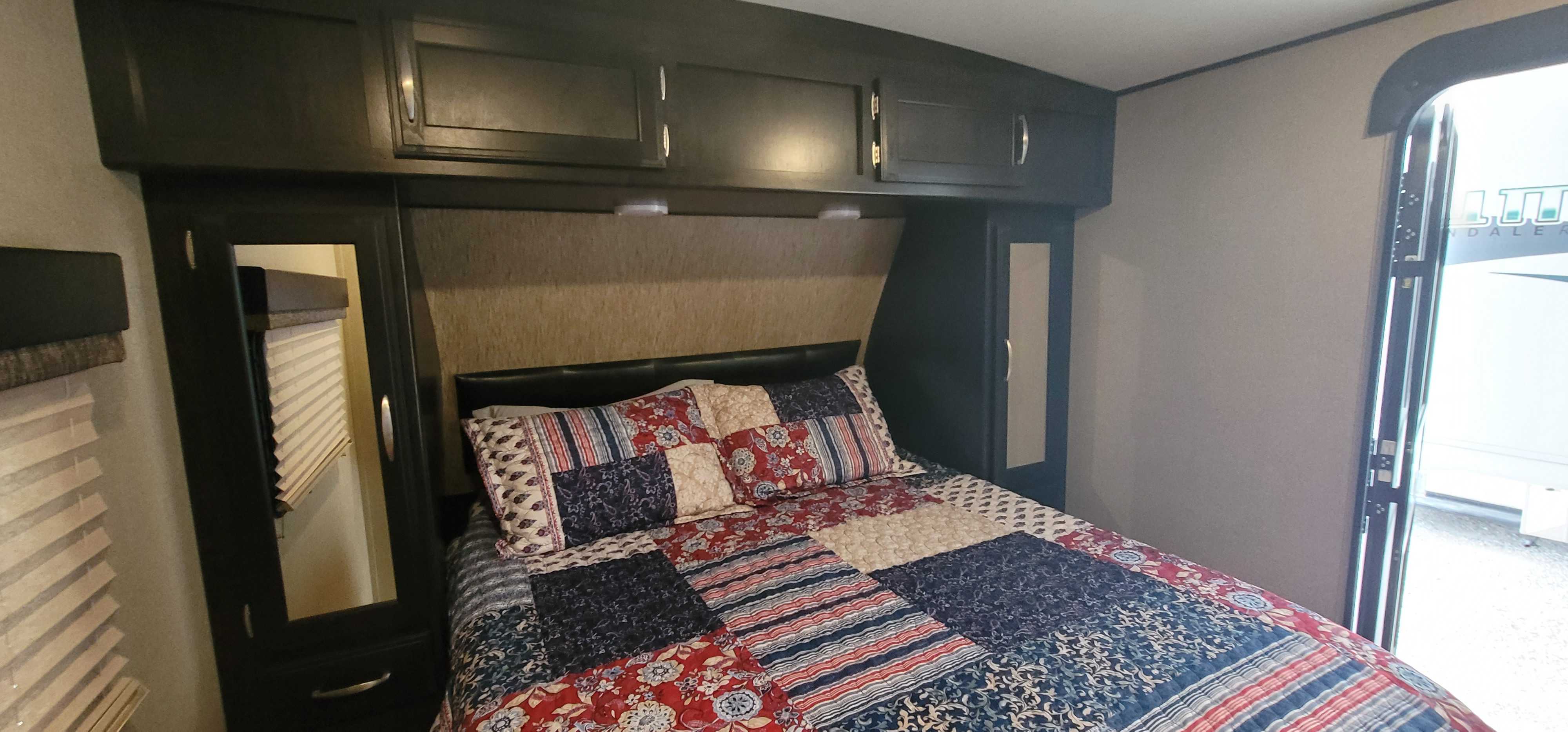 Cozy RV rental bedroom featuring a vibrant patchwork quilt, dark wood cabinets, and mirrored wardrobes for comfortable stays.. Grand Design Imagine 2019
