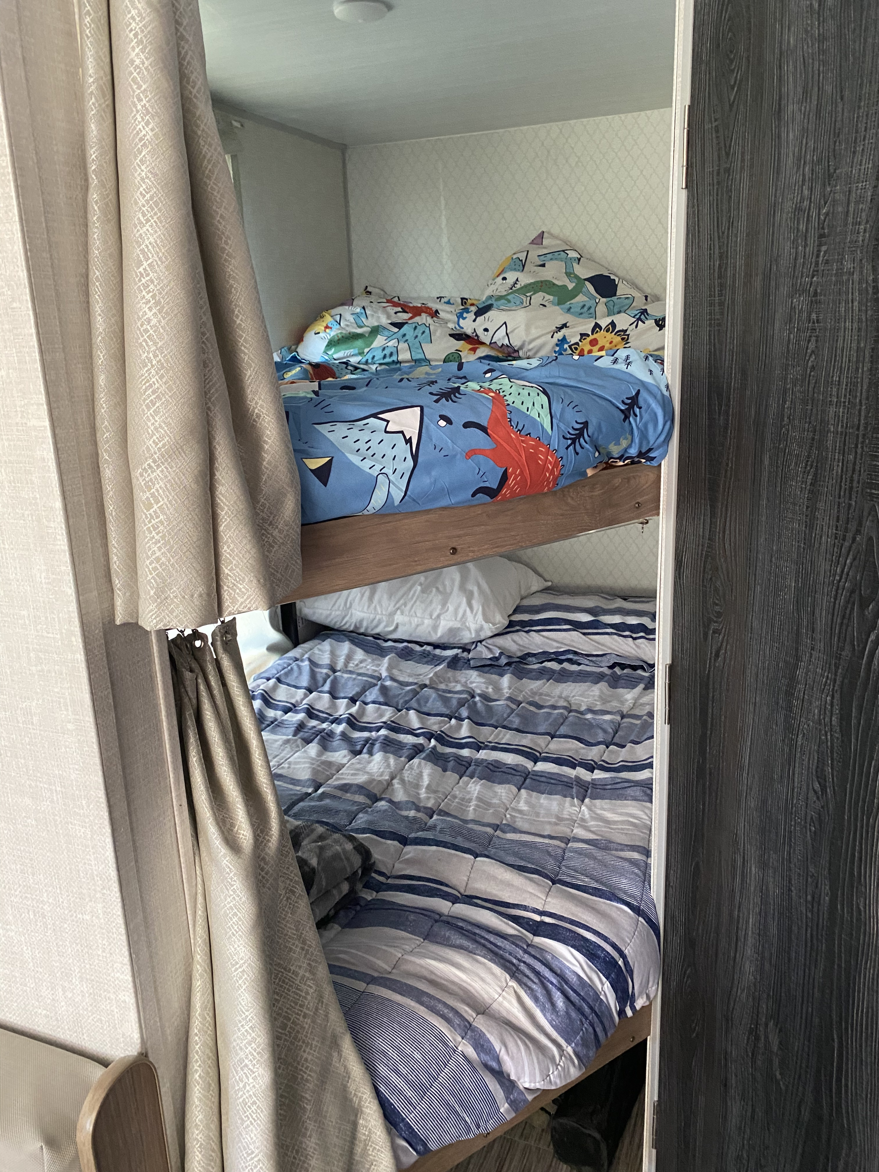 Inside an RV rental, showing cozy bunk beds: a top bunk with dinosaur bedding and a bottom bunk with blue striped bedding.. Forest River No Boundaries 19.3 2021