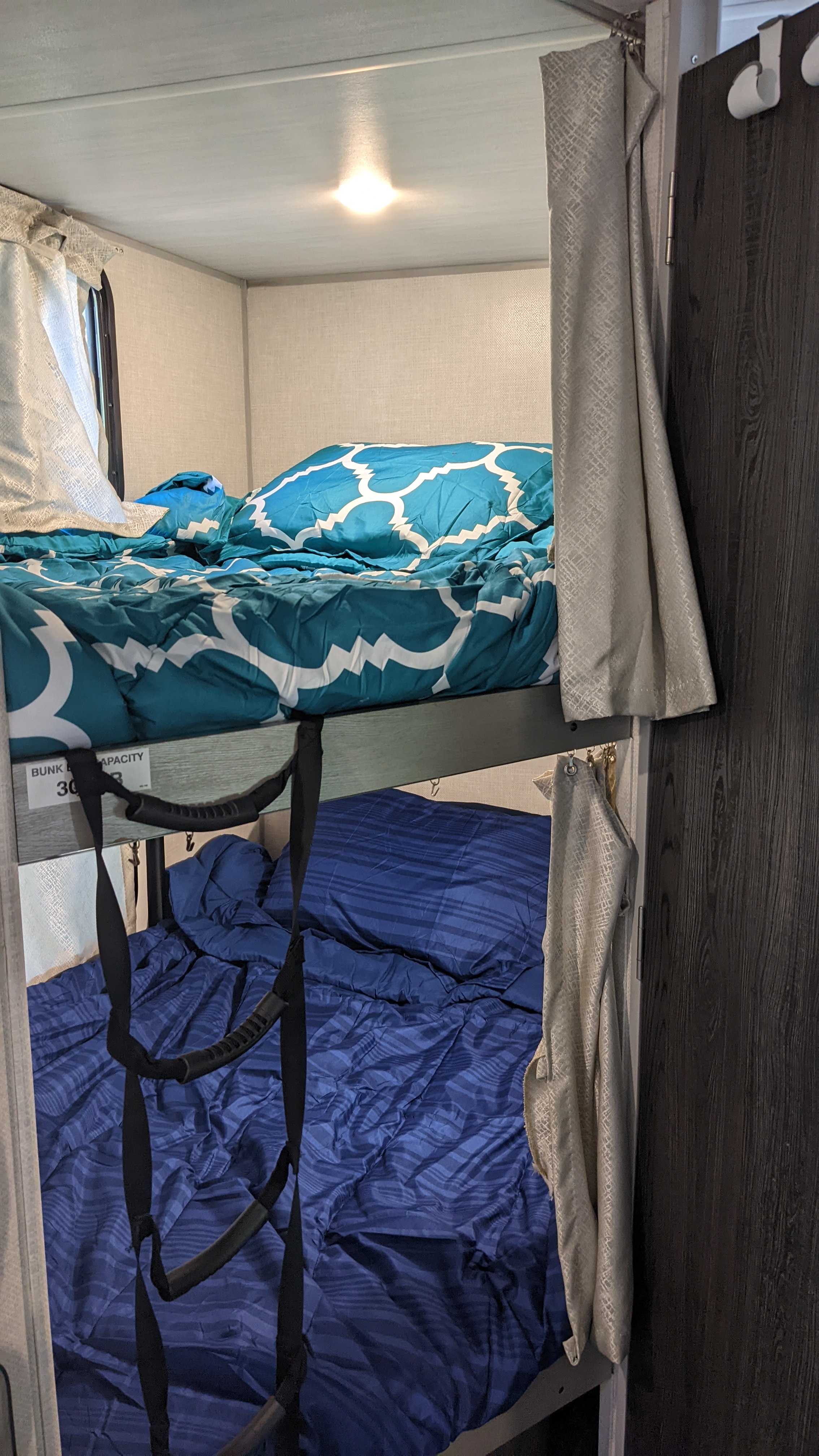An RV rental interior shows cozy bunk beds with colorful bedding (teal on top, blue below). A strap ladder hangs.. Forest River Ibex 2021