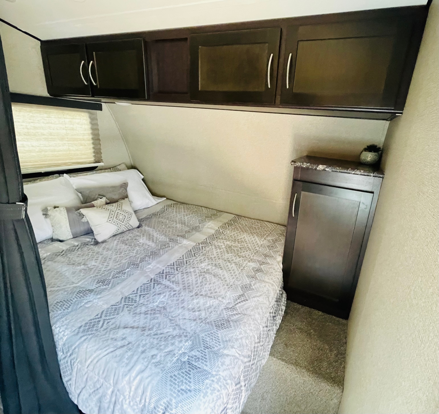 Comfortable RV rental bedroom: features a patterned bed, dark overhead cabinets, and a small side cabinet/nightstand.. Keystone Carbon 2017
