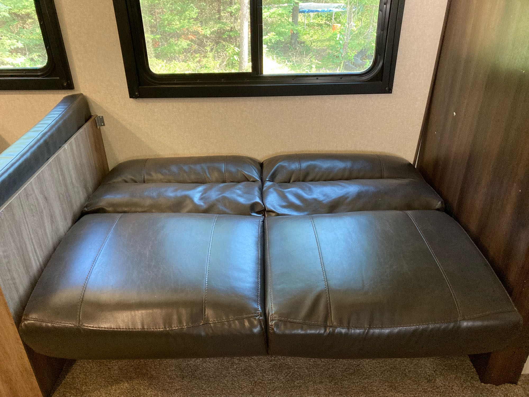 RV rental interior: A dark brown leather-like sofa bed, flattened for sleeping, under windows showing green trees.. Jayco Jay Feather 2018