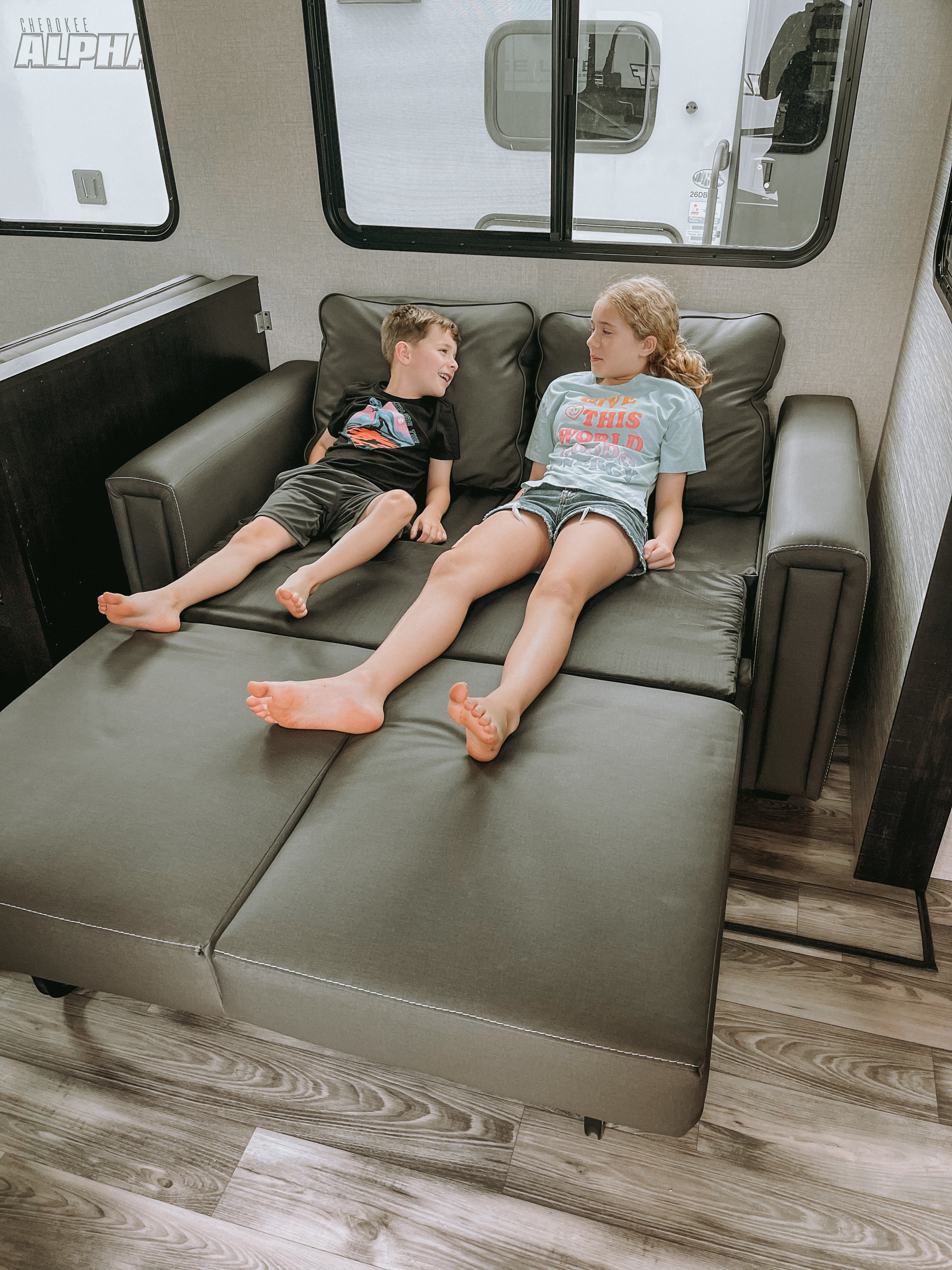 Two kids relax barefoot on a grey convertible sofa in an RV, smiling. Perfect for family travel & comfort in your rv rental.. Highland Ridge RV Range Lite 2022