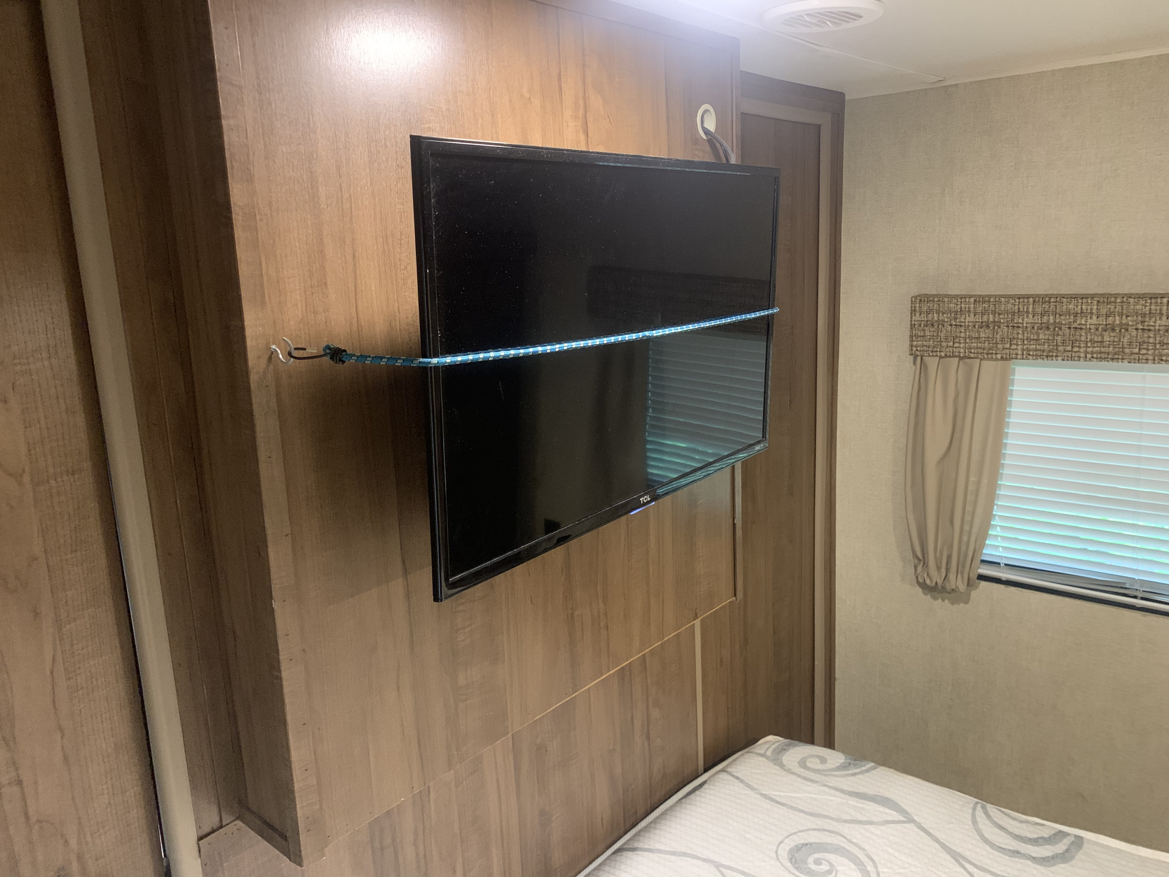 RV rental interior with a wall-mounted TV on wood paneling, secured by a blue bungee cord for travel. A bed and window are visible.. Gulf Stream Conquest 2019