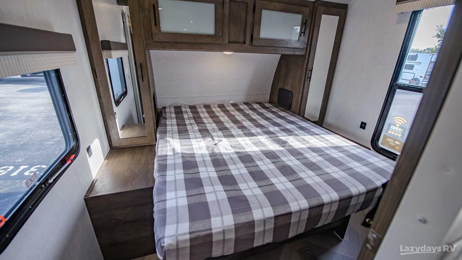 This cozy RV rental bedroom features a plaid bed, overhead cabinets, and large windows, offering comfortable travel accommodations.. Forest River Wildwood 2022