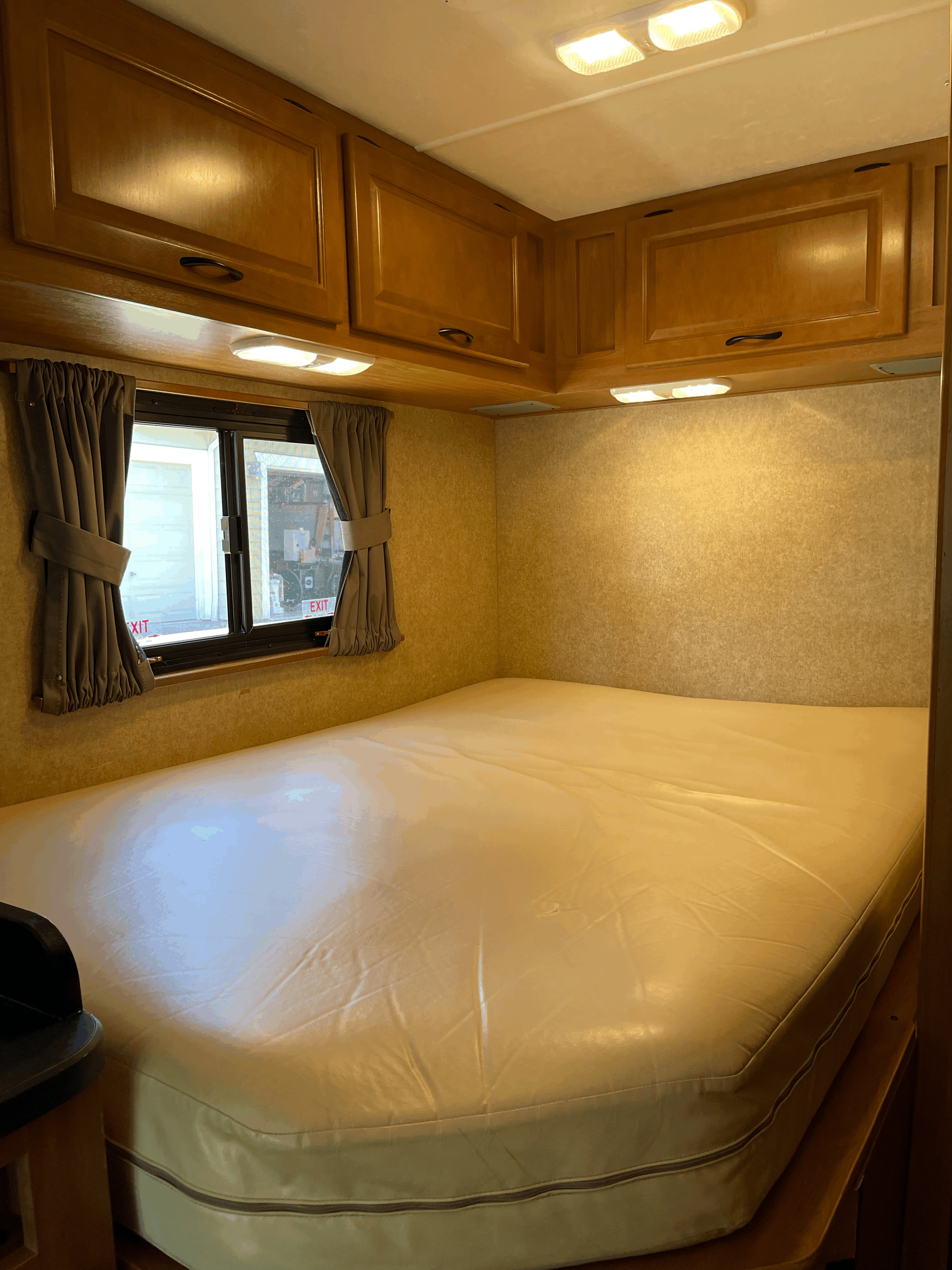 Spacious RV rental bedroom featuring a large mattress, overhead wood cabinets, and a window with drawn curtains.. Thor Majestic 2017