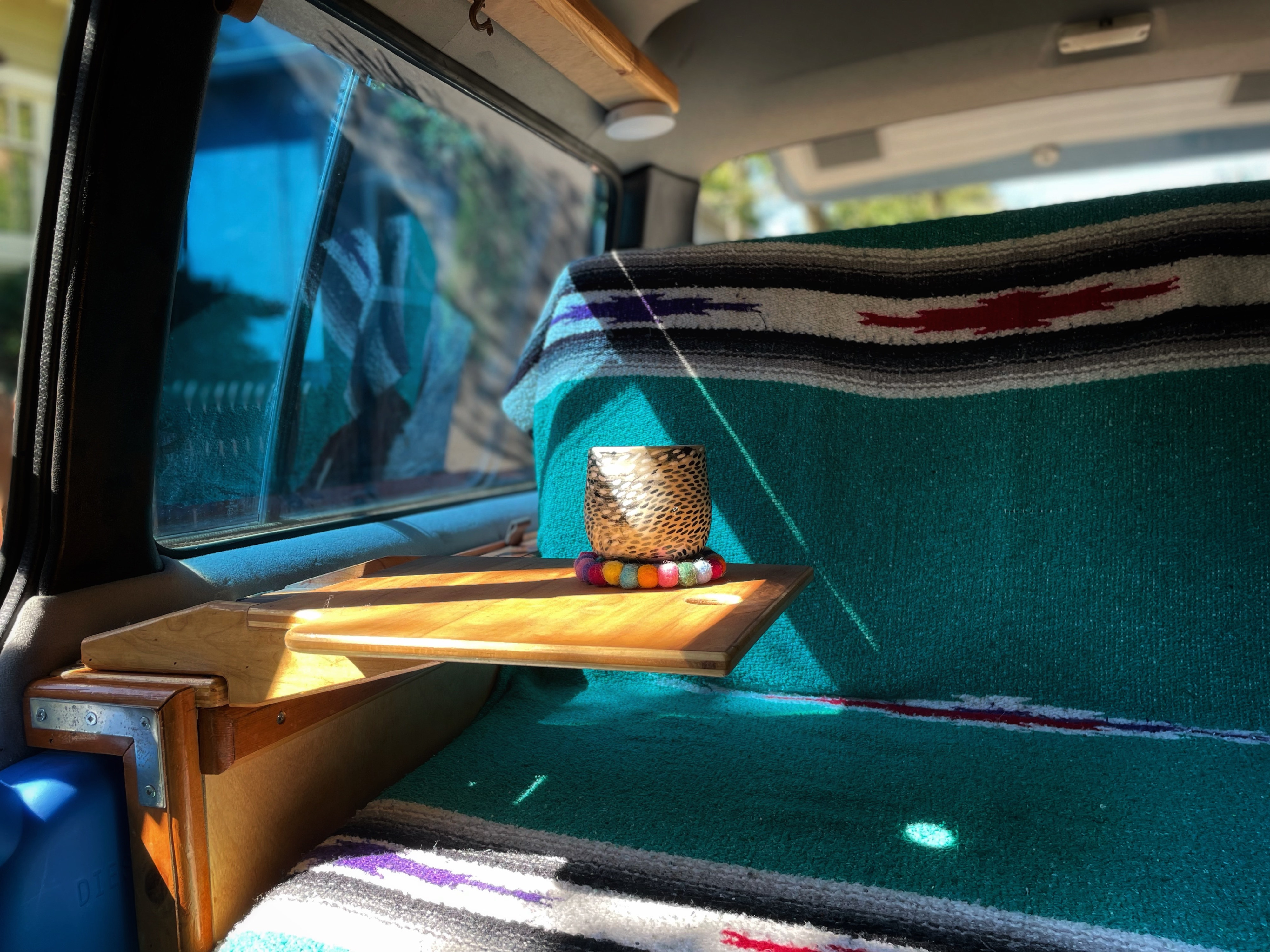 Enjoy the cozy RV rental interior, featuring a wooden shelf, patterned teal blanket, and a cup in sunlight.. Toyota Hiace 1985