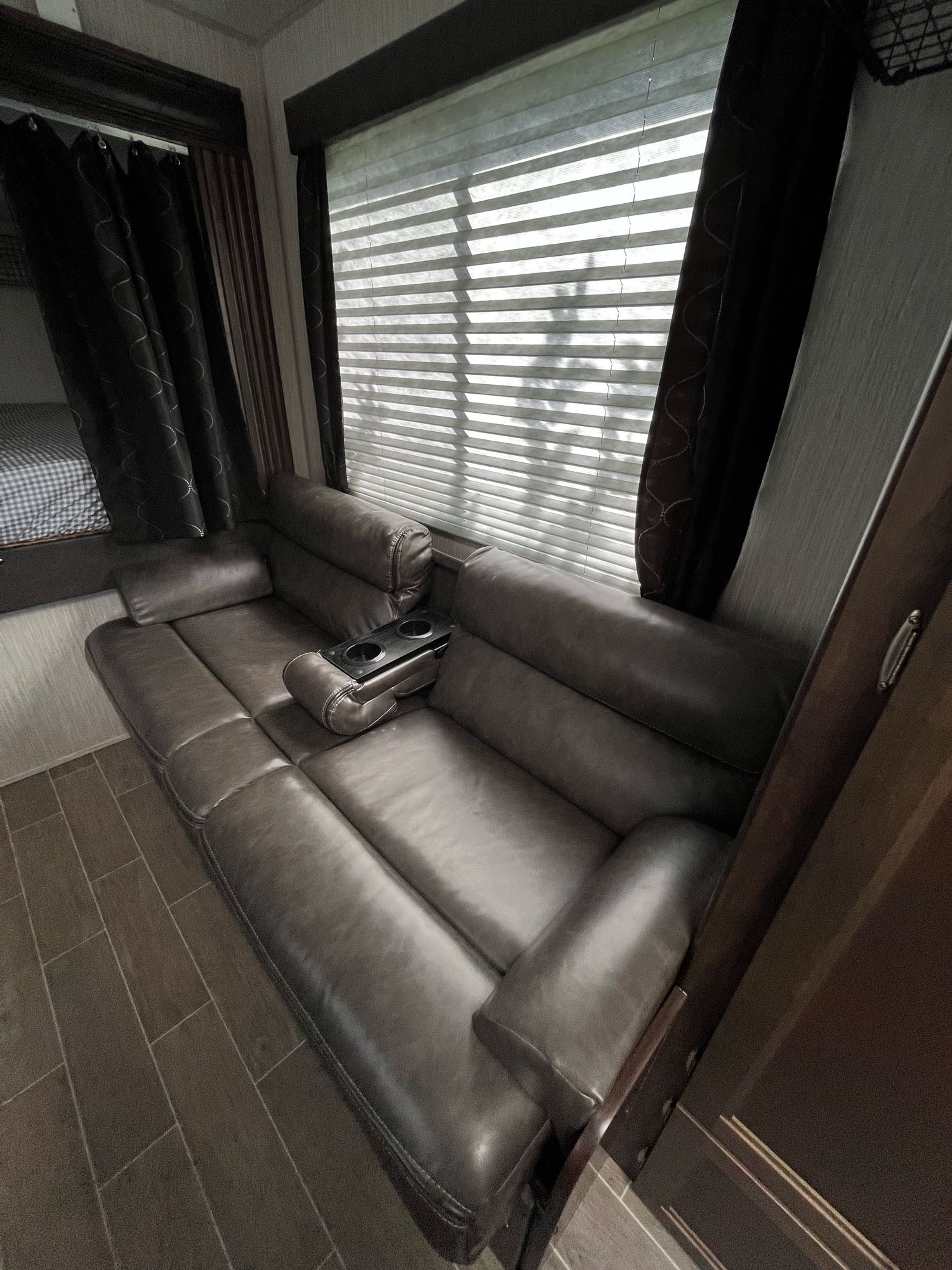 This RV rental features a comfortable, dark brown leather reclining sofa with cup holders, alongside a window with blinds revealing sunlight.. Forest River Cherokee Grey Wolf 2020