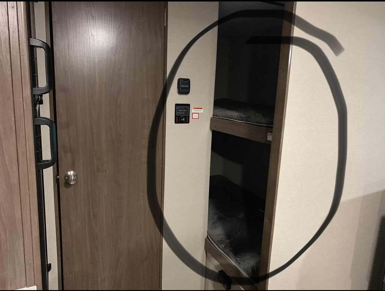 RV rental interior: a light wood door, wall controls, and cozy bunk beds with dark mattresses, framed by a dark circle.. Keystone RV Hideout 2019