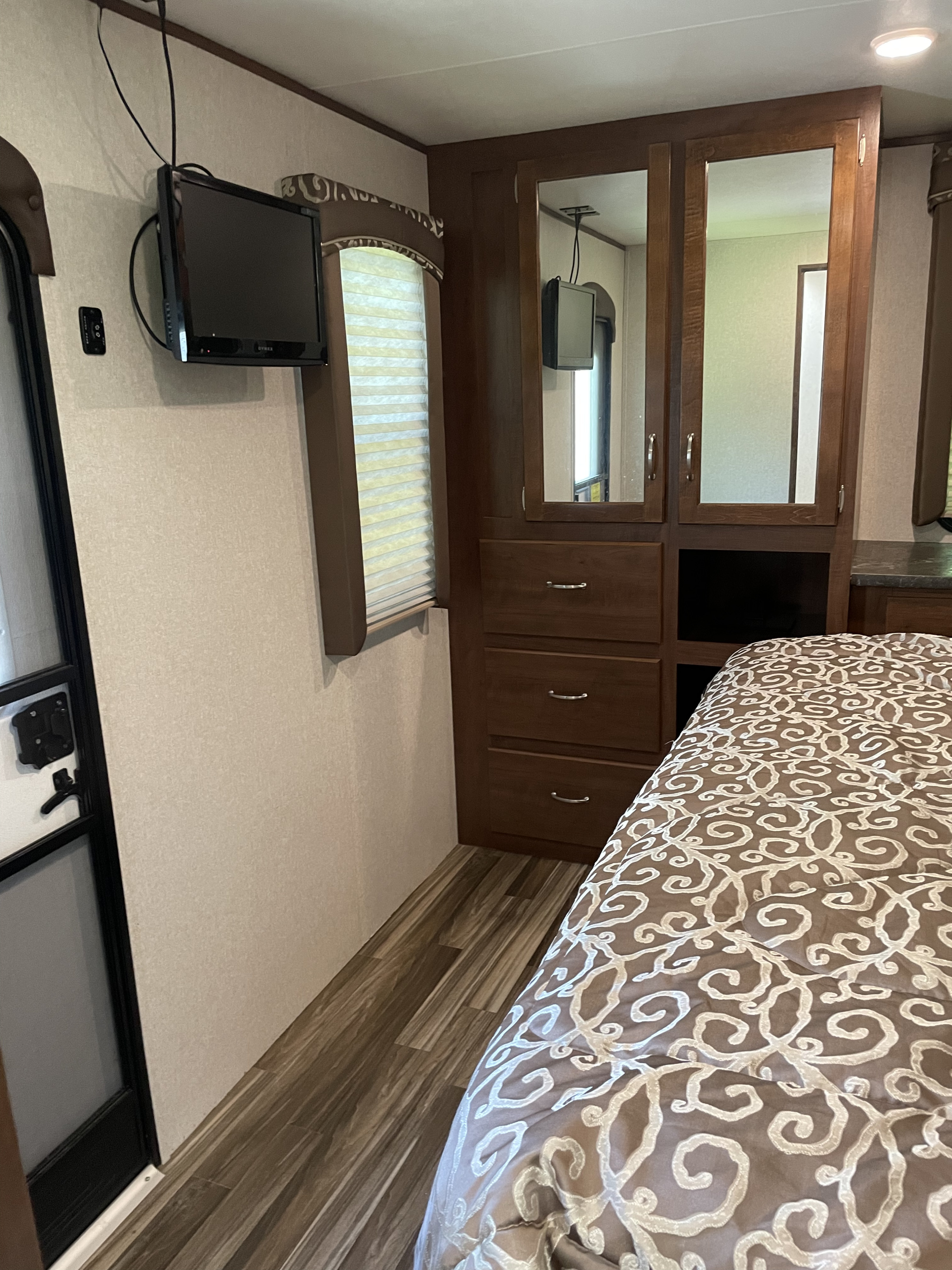 An RV rental bedroom features a wall-mounted TV, built-in wardrobe with drawers, a bed with a patterned comforter, and blinds.. Jayco Jay Flight 2018