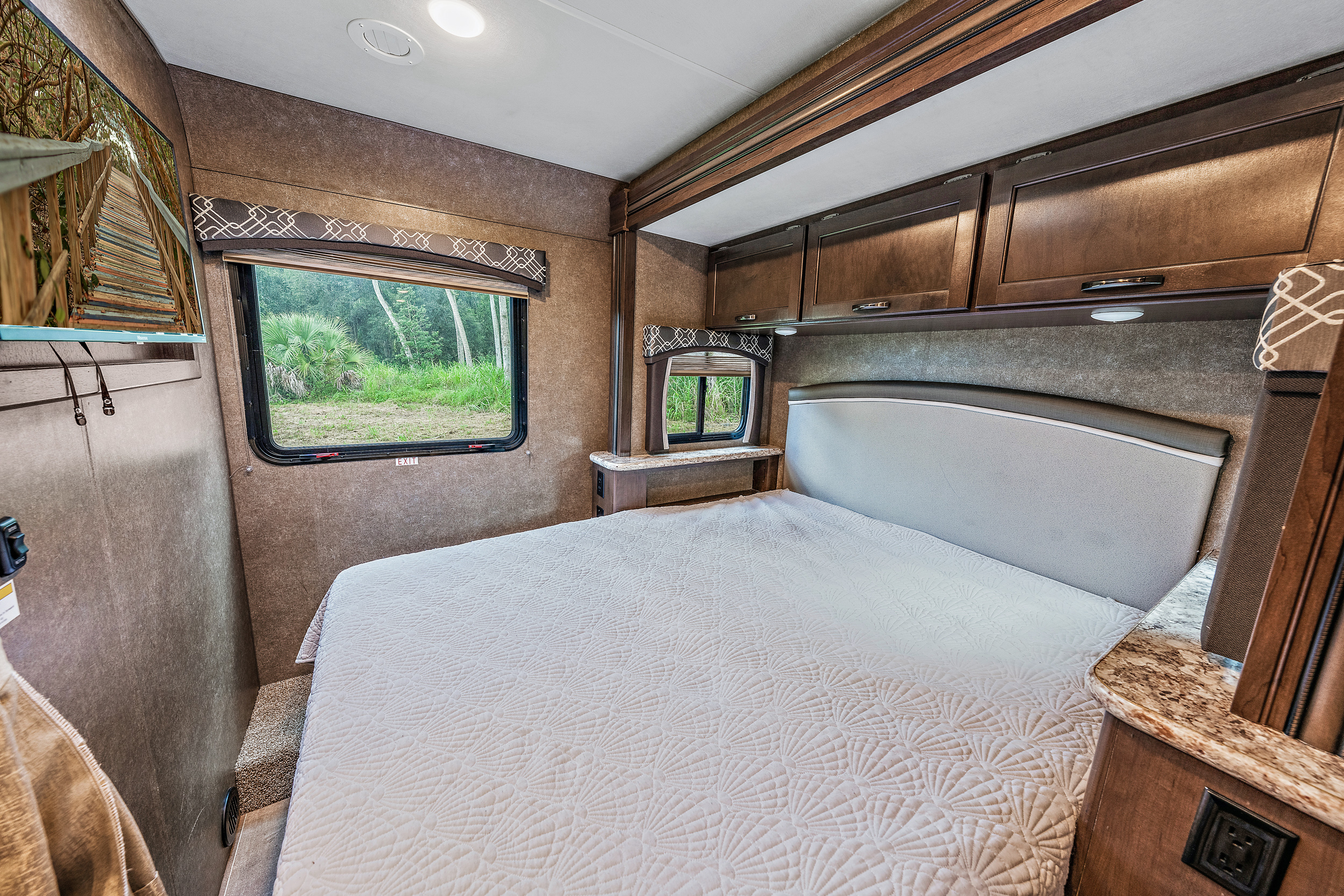 Cozy RV rental bedroom with a comfortable bed, wood cabinets, wall-mounted TV, and a nature view from the windows.. Thor Motor Coach Chateau 2020