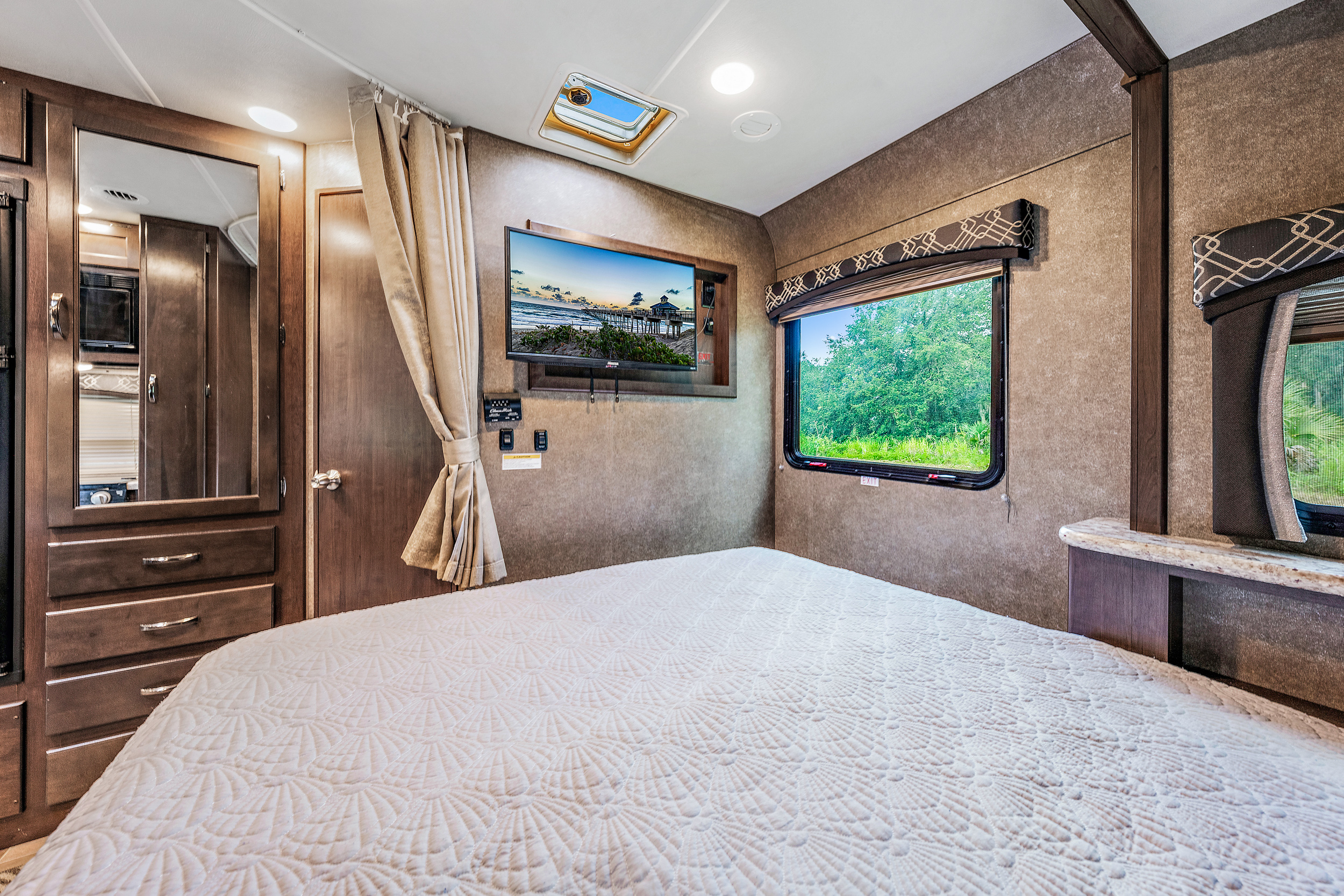 Experience comfort in this RV rental bedroom, featuring a TV, plush bed, cabinetry, and a window with nature views.. Thor Motor Coach Chateau 2020