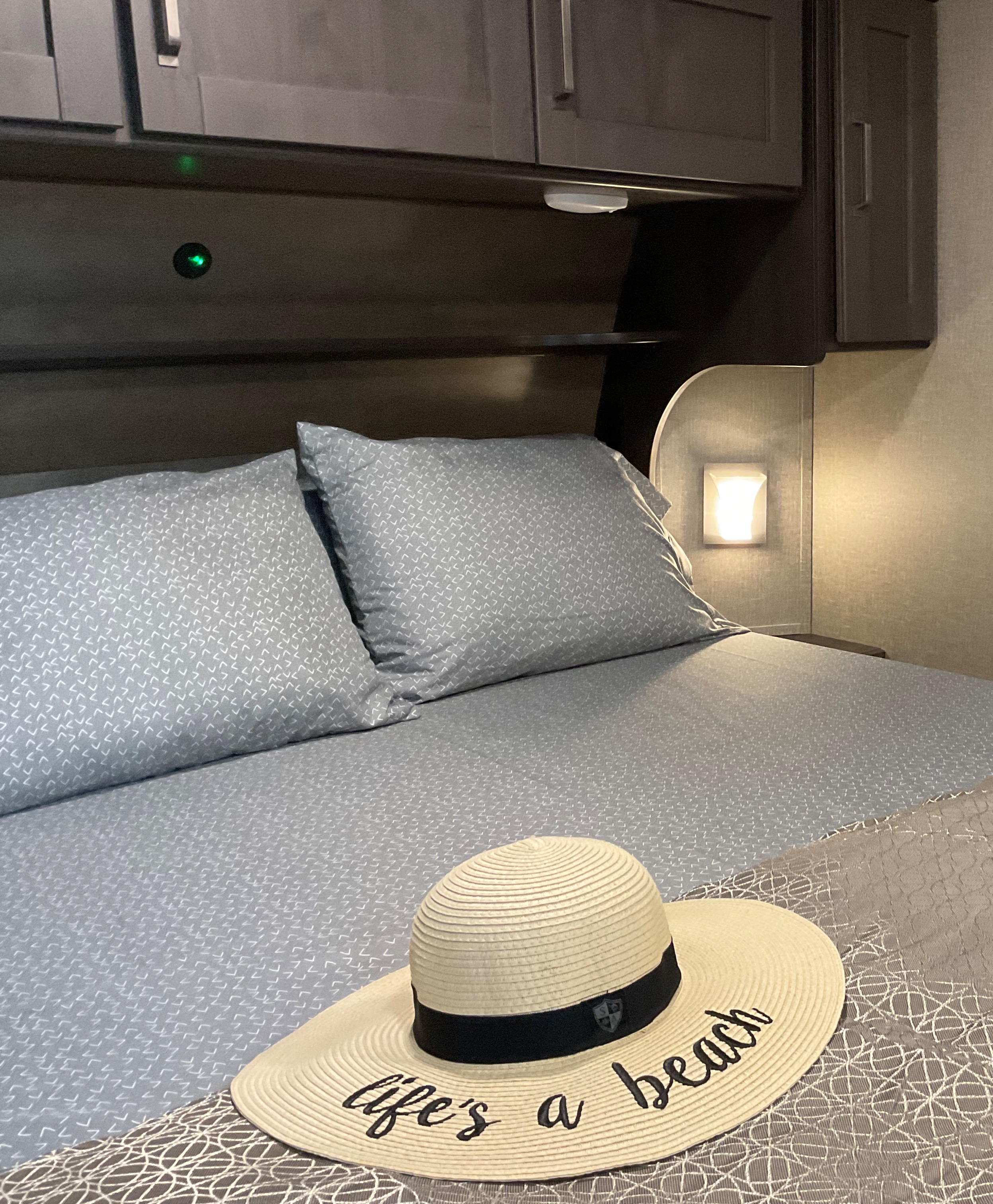 Relax in your cozy RV rental! This inviting bedroom features patterned bedding, soft pillows, and a sun hat with "life's a beach" on it.. Grand Design Reflection RLS (Rear Living Area) 2022
