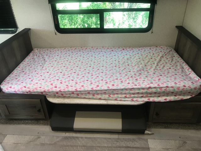 Cozy RV rental bed with heart-patterned sheet, a large window offering green views, and practical under-bed storage units.. KZ Sportsmen LE 2021