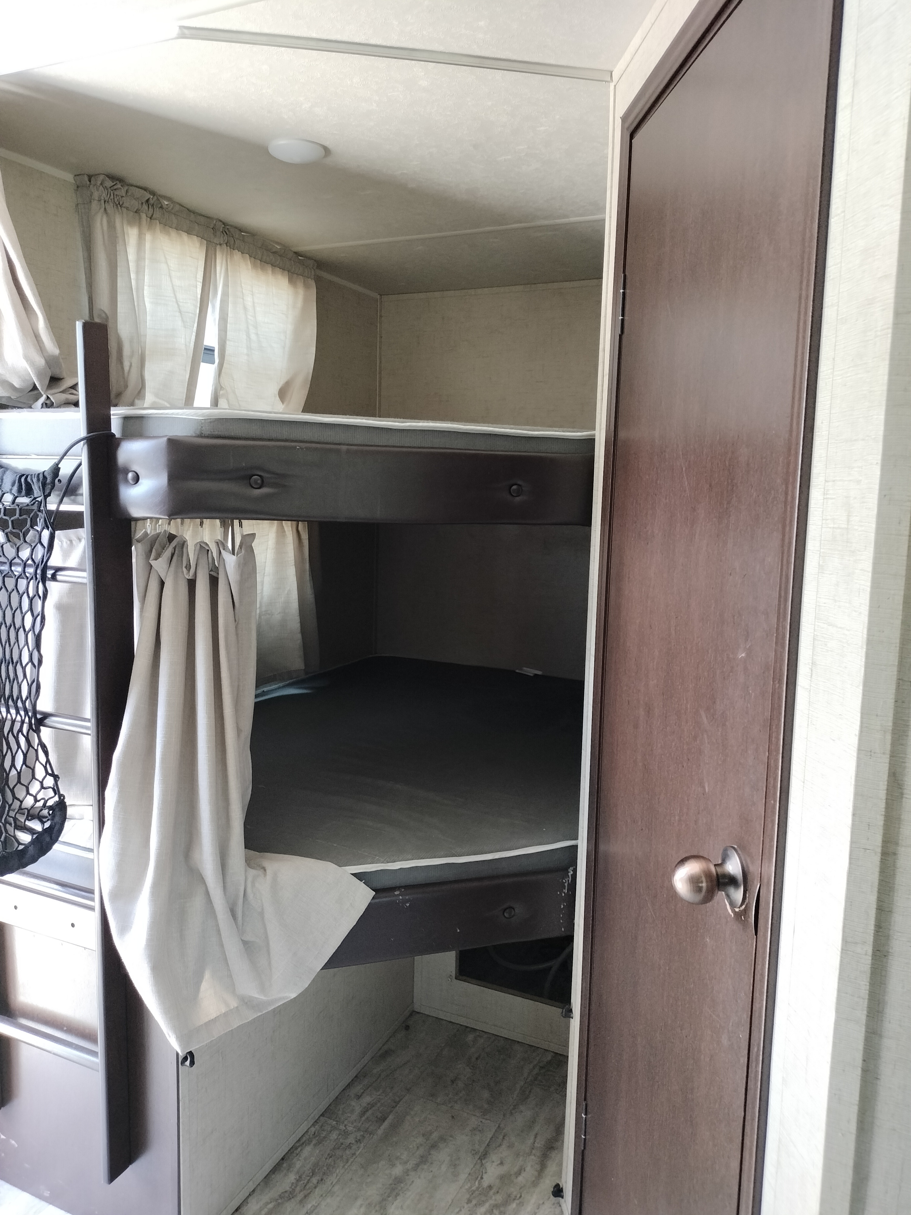 Inside an RV rental: two compact bunk beds with mattresses, light curtains, and a brown door, optimizing sleeping space.. East to West Della Terra 2020