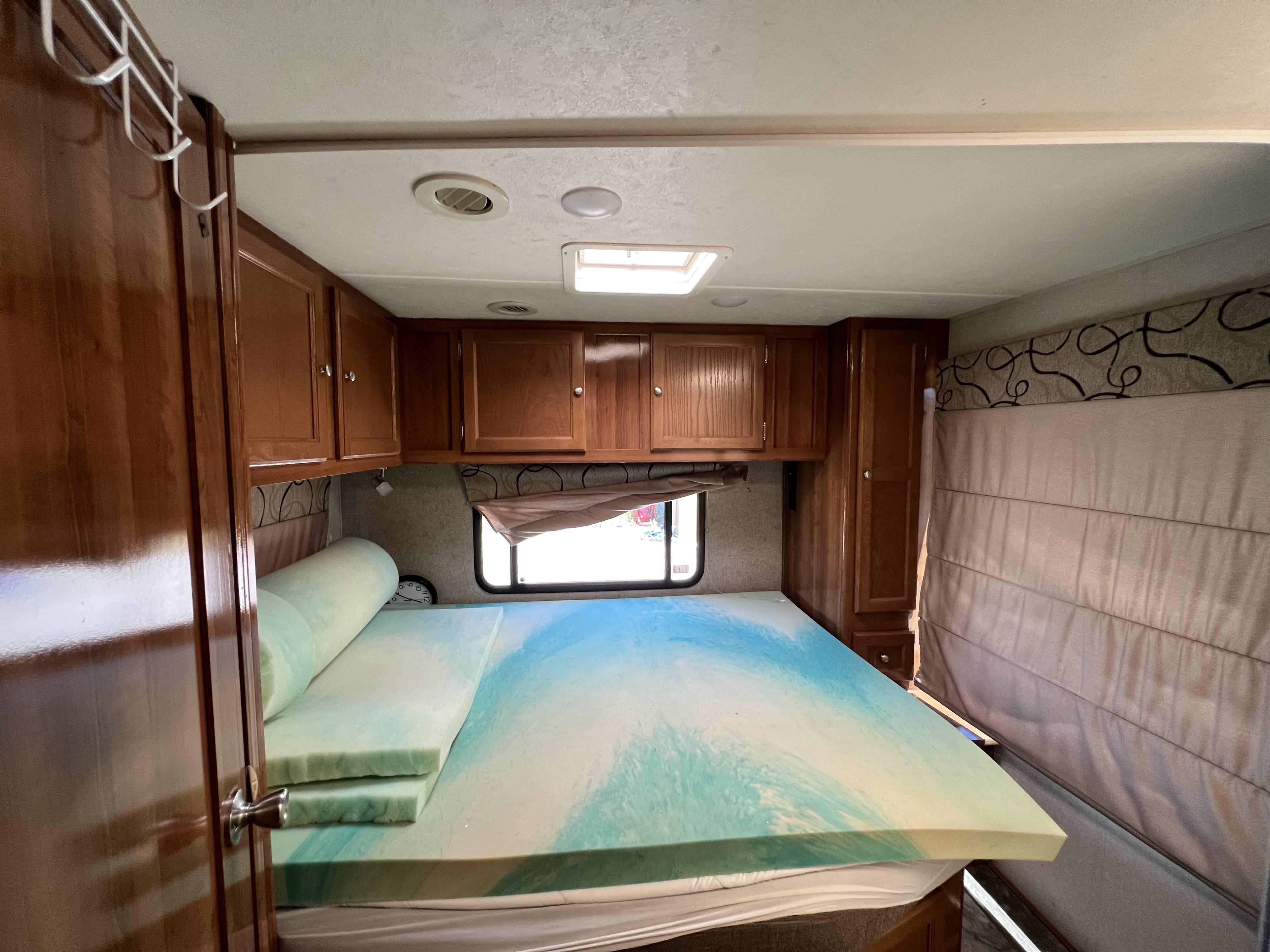 RV rental bedroom showcasing a bed with a layered foam mattress topper, wooden cabinets, and a window for natural light.. Coachmen Leprechaun 2019