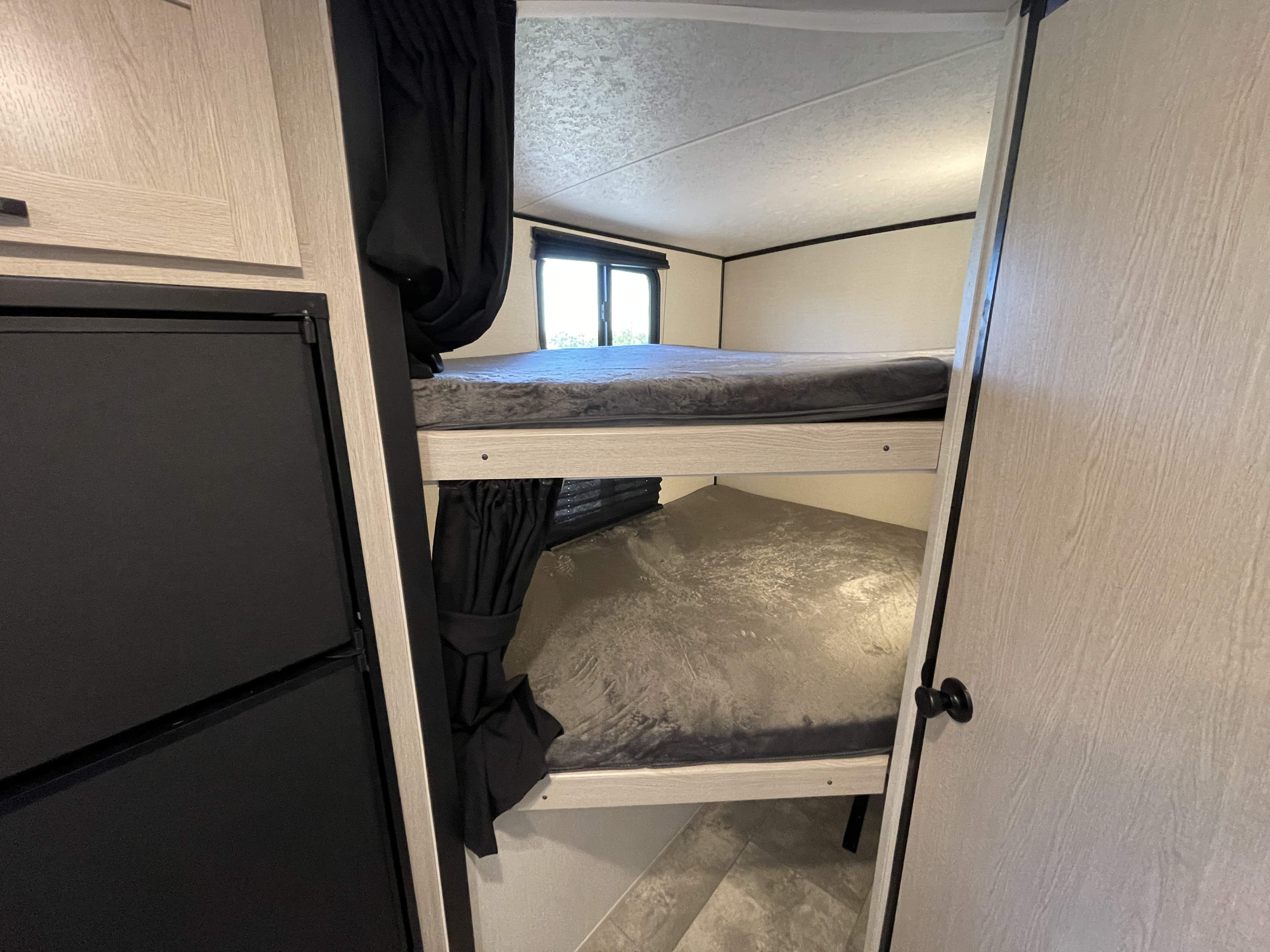 Cozy RV rental bunk beds. Interior view showing grey-bedded upper and lower bunks, a black refrigerator, and light wood cabinetry.. Coachmen Apex 2022
