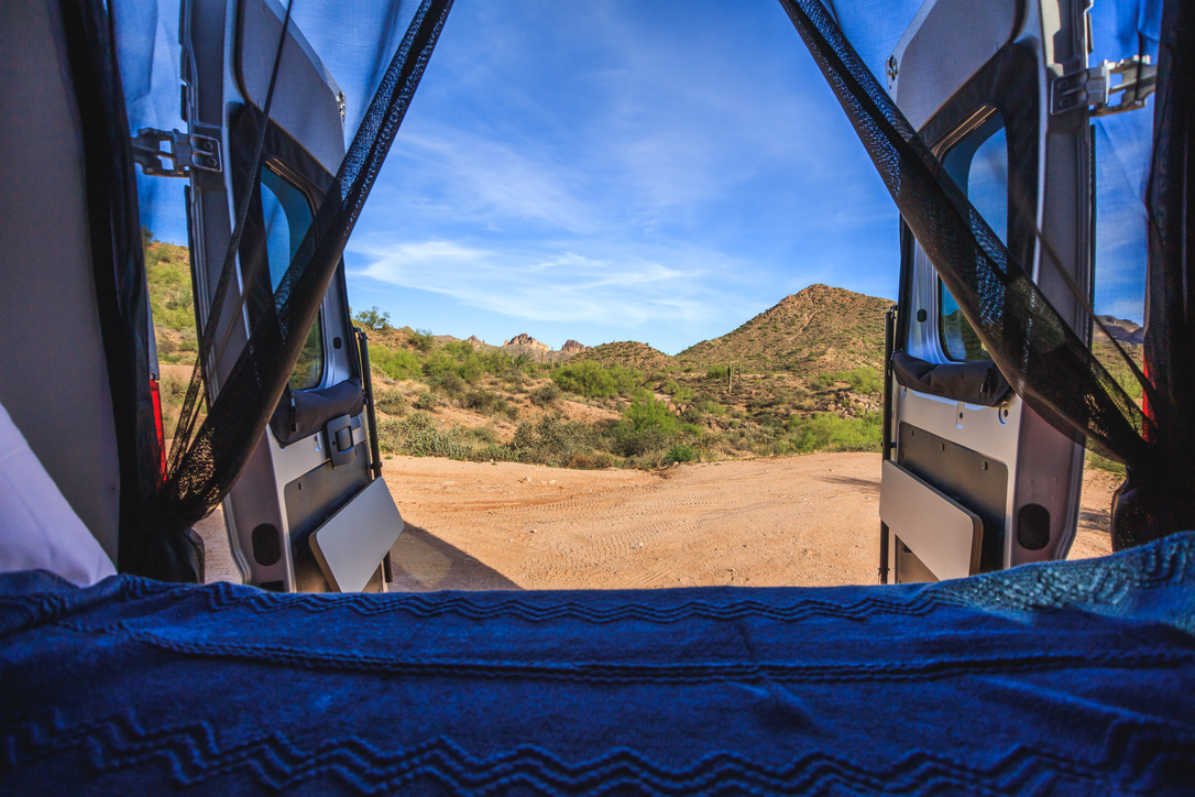 A cozy blue bed in an RV rental looks out open camper van doors to a sunny desert landscape with hills.. Winnebago Solis 2022
