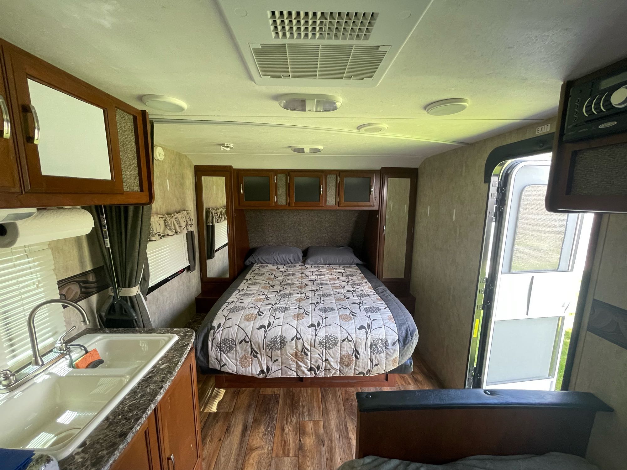 This RV rental interior features a cozy bedroom with a floral bedspread, a small kitchen sink, and wood flooring.. Keystone RV Passport Ultra Lite 2015