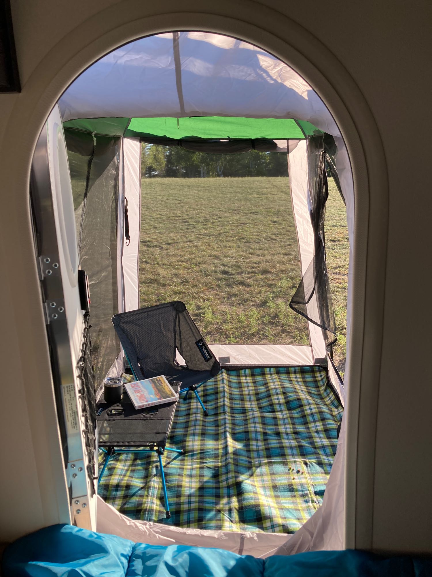 View from an rv rental, looking into an attached screen room setup with a folding chair, table, plaid mat, and grassy field.. Nucamp T@G 2022