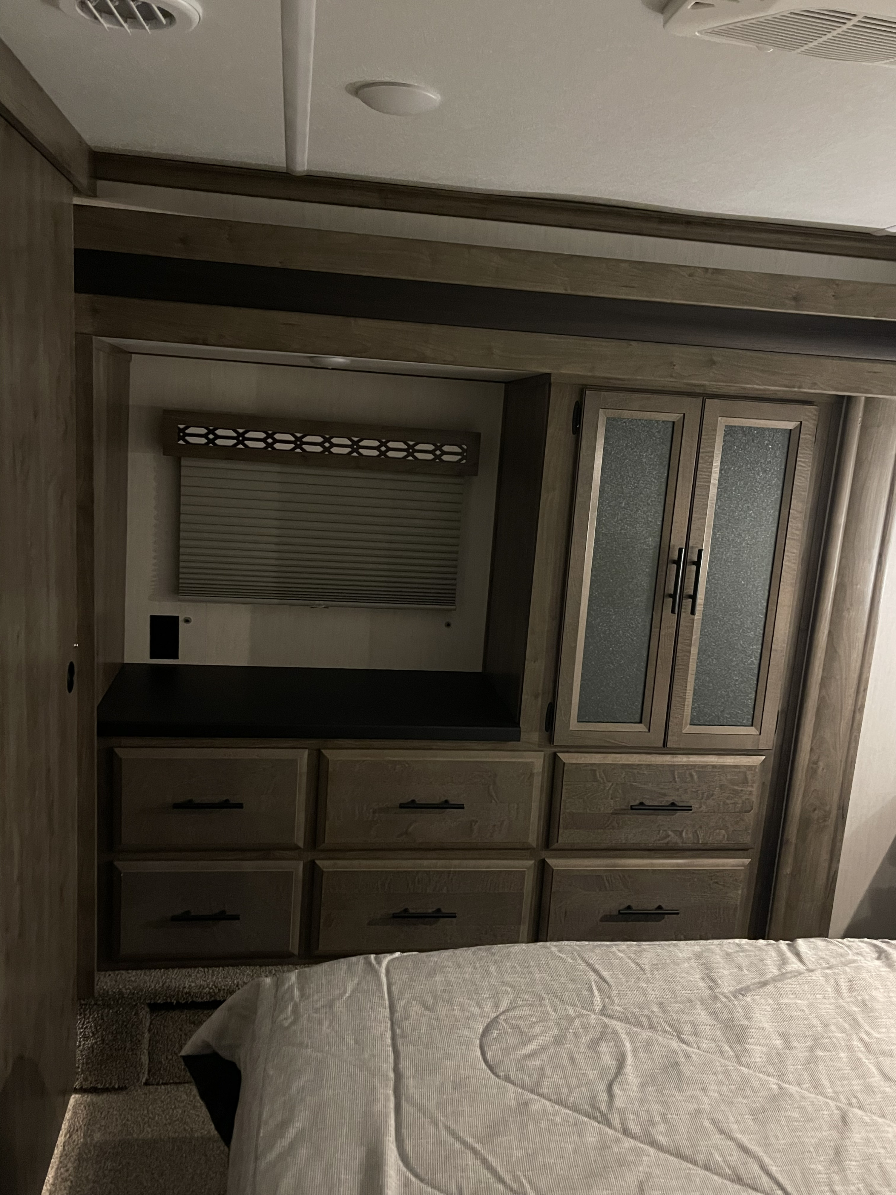An RV rental bedroom showing a bed, built-in wooden storage with drawers, a wardrobe with frosted doors, and a window blind.. Forest River Vengeance 2021