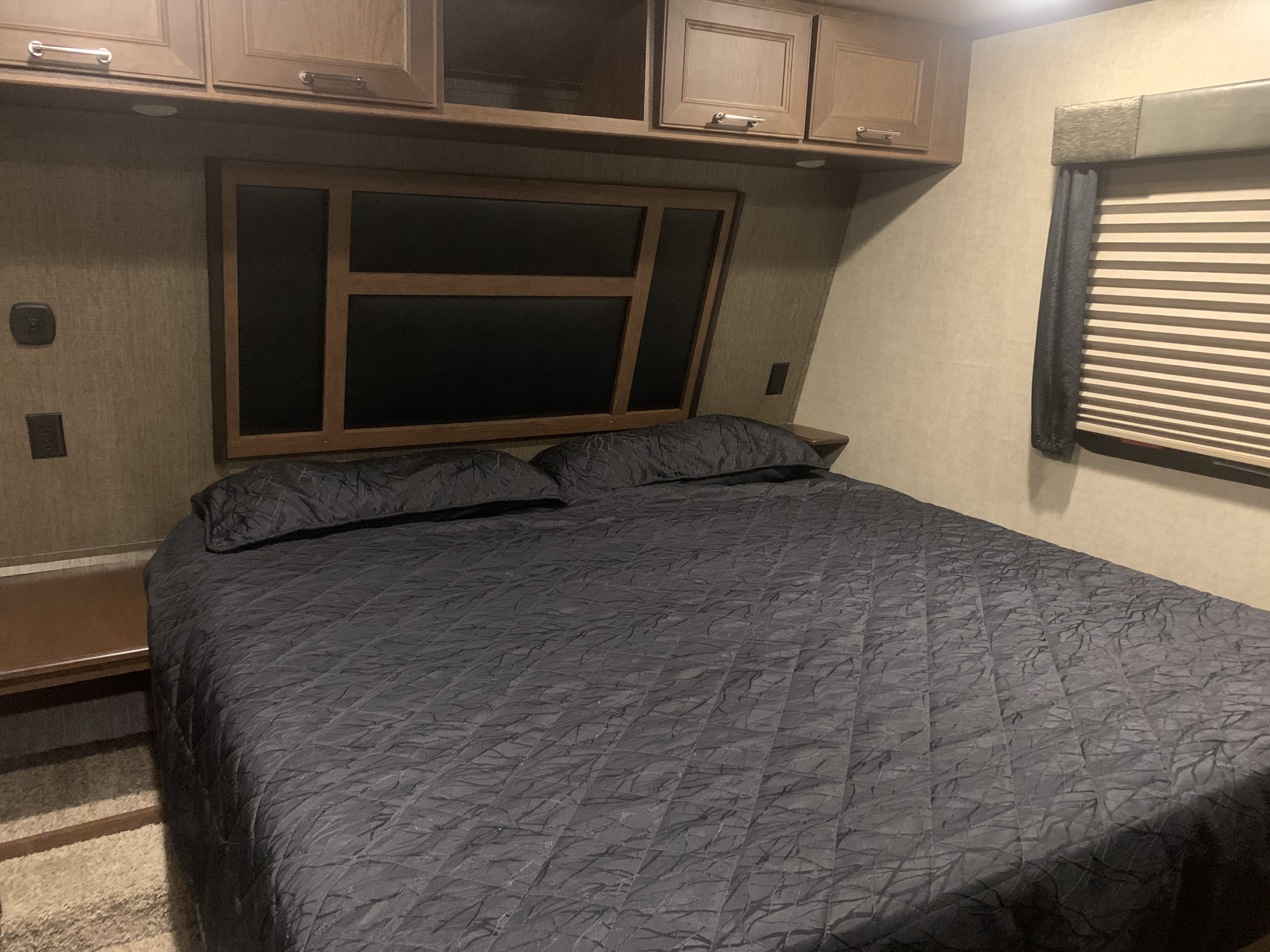 Comfortable RV rental bedroom featuring a large bed with dark bedding, overhead cabinets, and a window with blinds.. Keystone Fuzion 2015