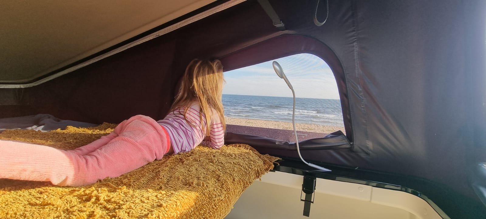 Cozy RV rental view! A child relaxes on a bed, looking out at the calm ocean and sandy beach from a pop-top camper.. Ford Randger R530 2022