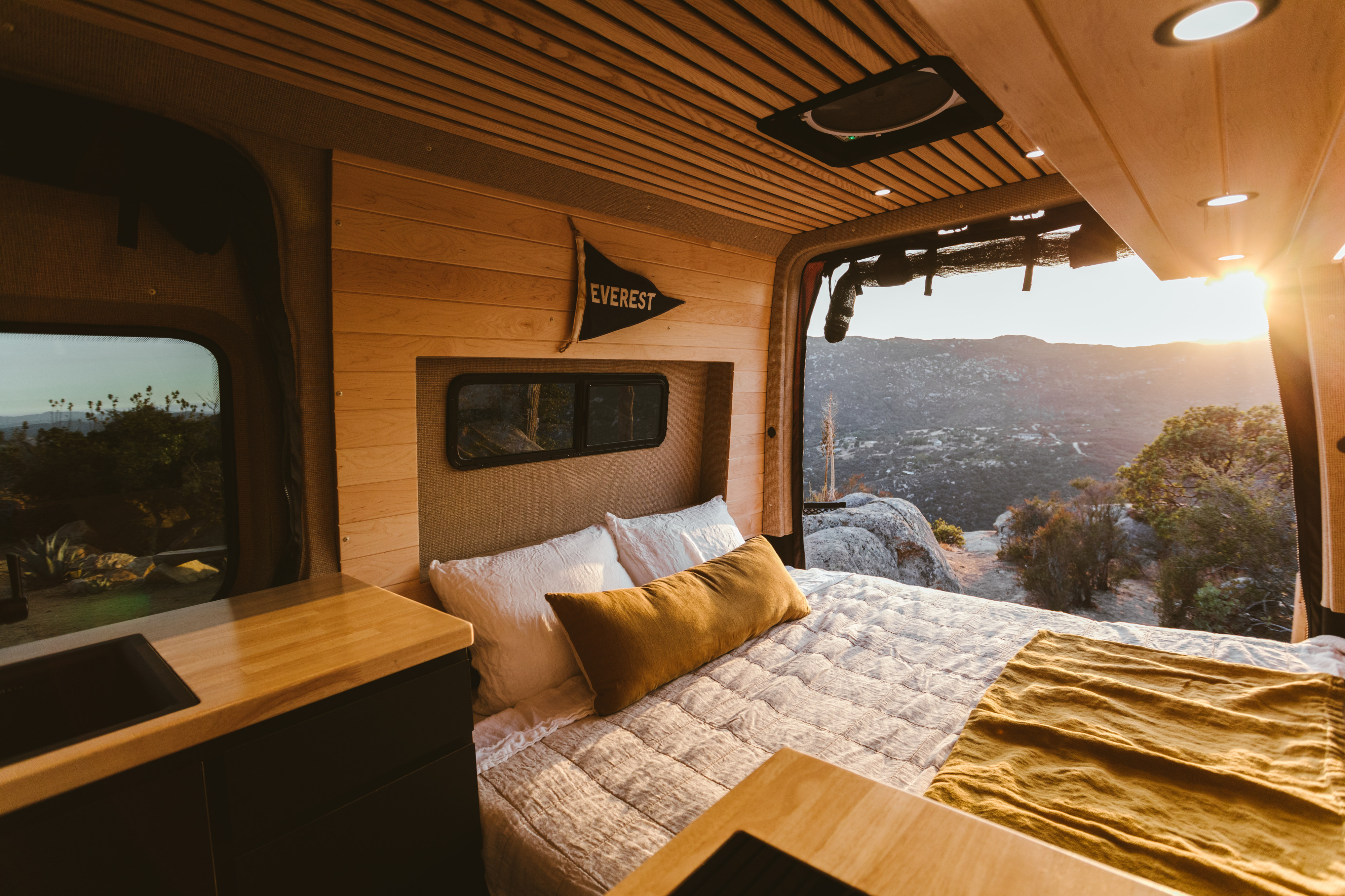 An RV rental's cozy bedroom features wood paneling & an open back, revealing a golden sunset over a rocky mountain landscape.. Mercedes-Benz Sprinter 2021