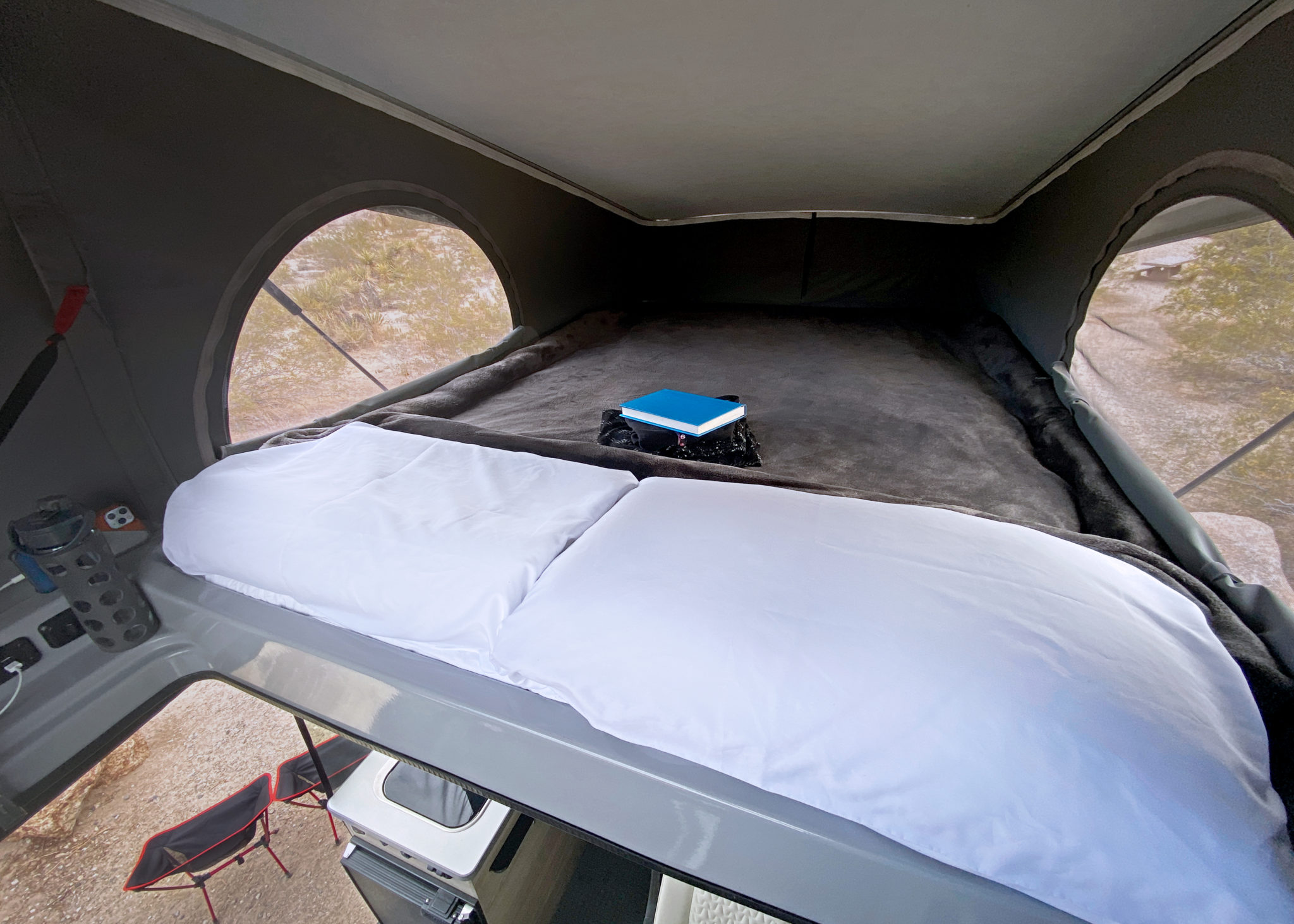 RV rental's elevated sleeping area: a comfy bed with white sheets, a blue book, and arched windows with an outdoor view.. Winnebago Solis 2021