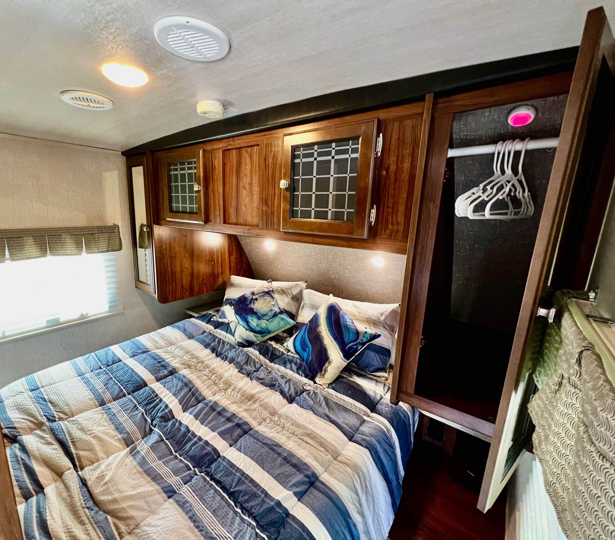 Cozy RV rental bedroom with striped bed, overhead cabinets, built-in closet, and window. Ideal for travel comfort.. Heartland RVs Mallard 2019