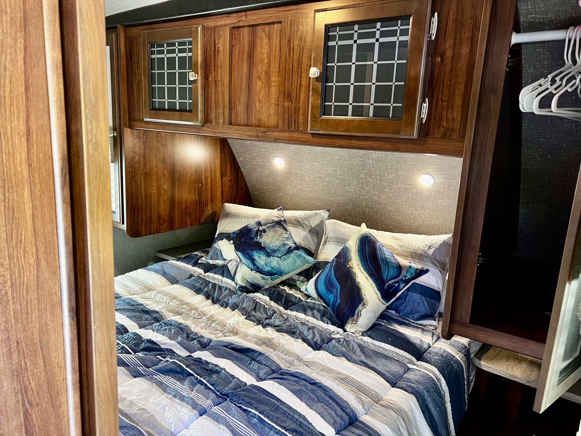 Comfortable RV rental bedroom featuring a striped bed, artistic pillows, overhead wood cabinets with glass inserts, and bedside reading lights.. Heartland RVs Mallard 2019