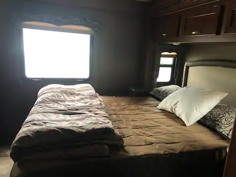 Cozy RV rental bedroom with a queen bed, brown bedding, two bright windows, and overhead cabinets.. Thor Four Winds 2017