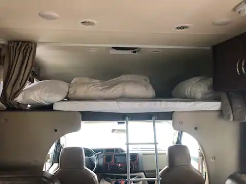 Spacious RV rental interior showing an overhead sleeping bunk with bedding and ladder leading to the front cockpit.. Thor Four Winds 2017