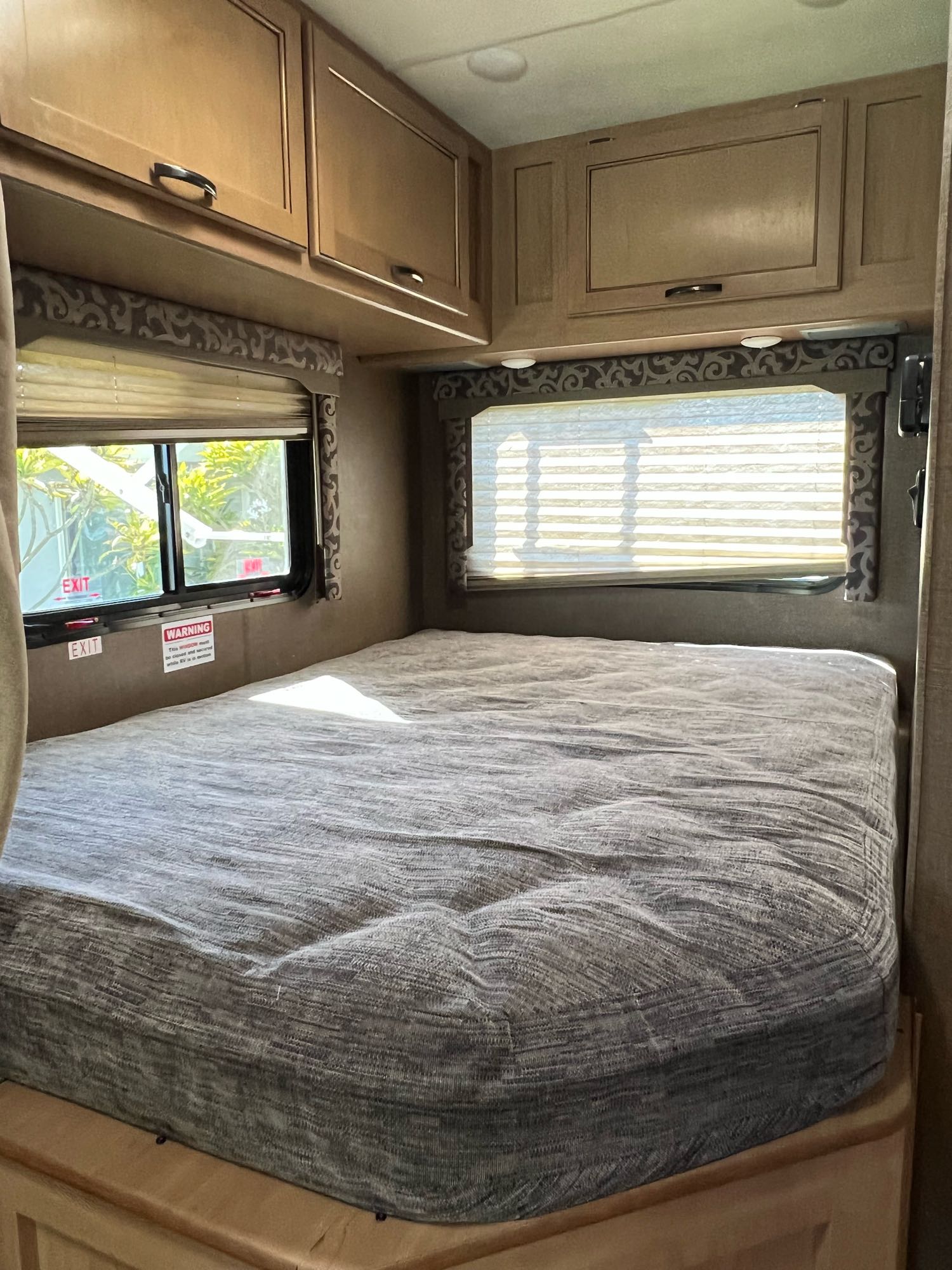 View of an RV rental bedroom with a gray mattress, wooden overhead cabinets, and two windows for light.. Thor Four Winds 2019