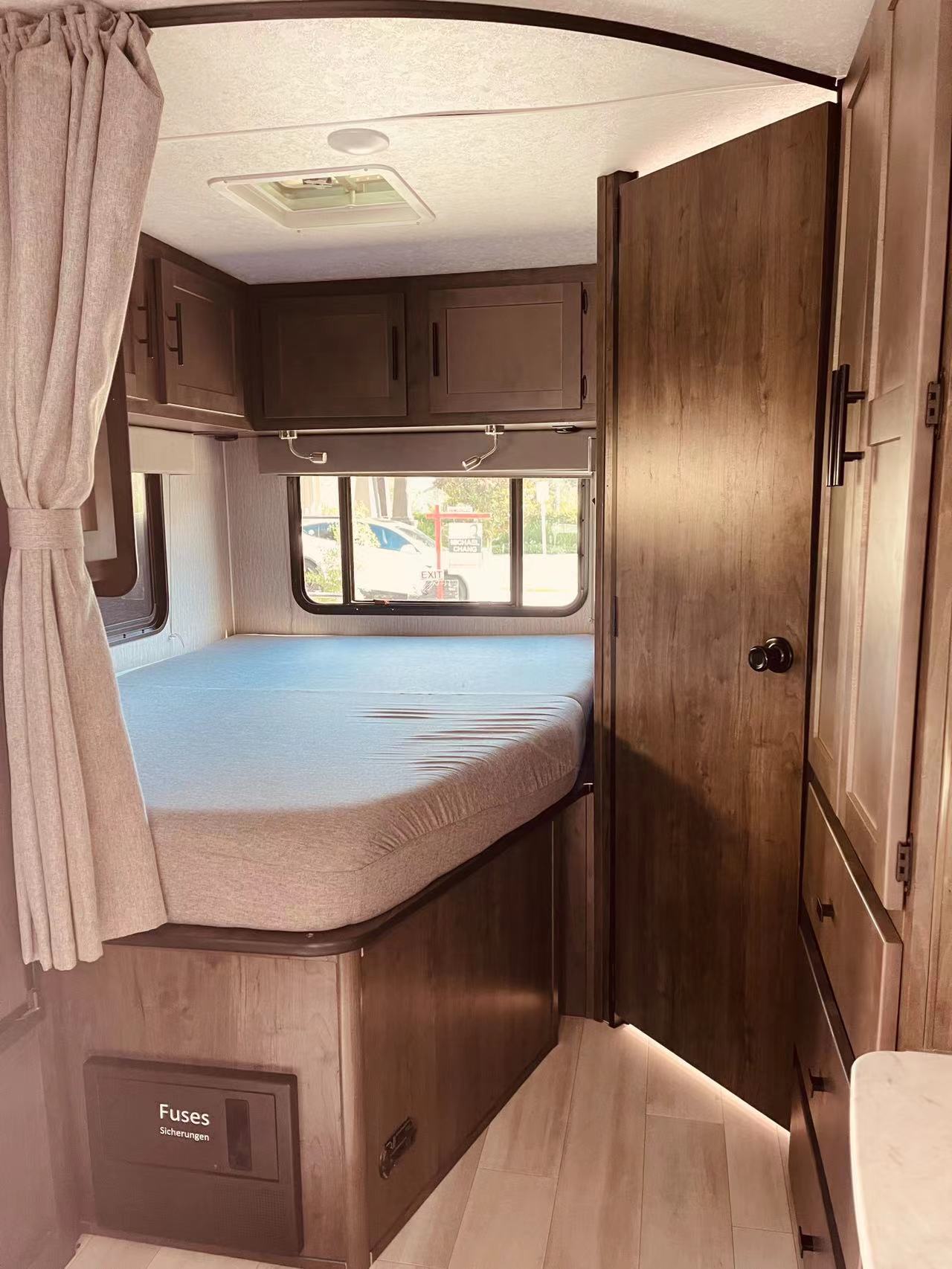 Inside an RV rental: a compact bedroom with a bed, overhead cabinets, a wardrobe, and a visible fuse box.. Forest River Coachmen Leprechaun 2022