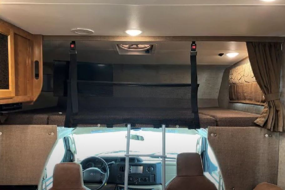 Inside an RV rental: a cozy cab-over bunk with safety net above the driver's cockpit, illuminated by interior lighting.. Forest River Coachmen Leprechaun 2018