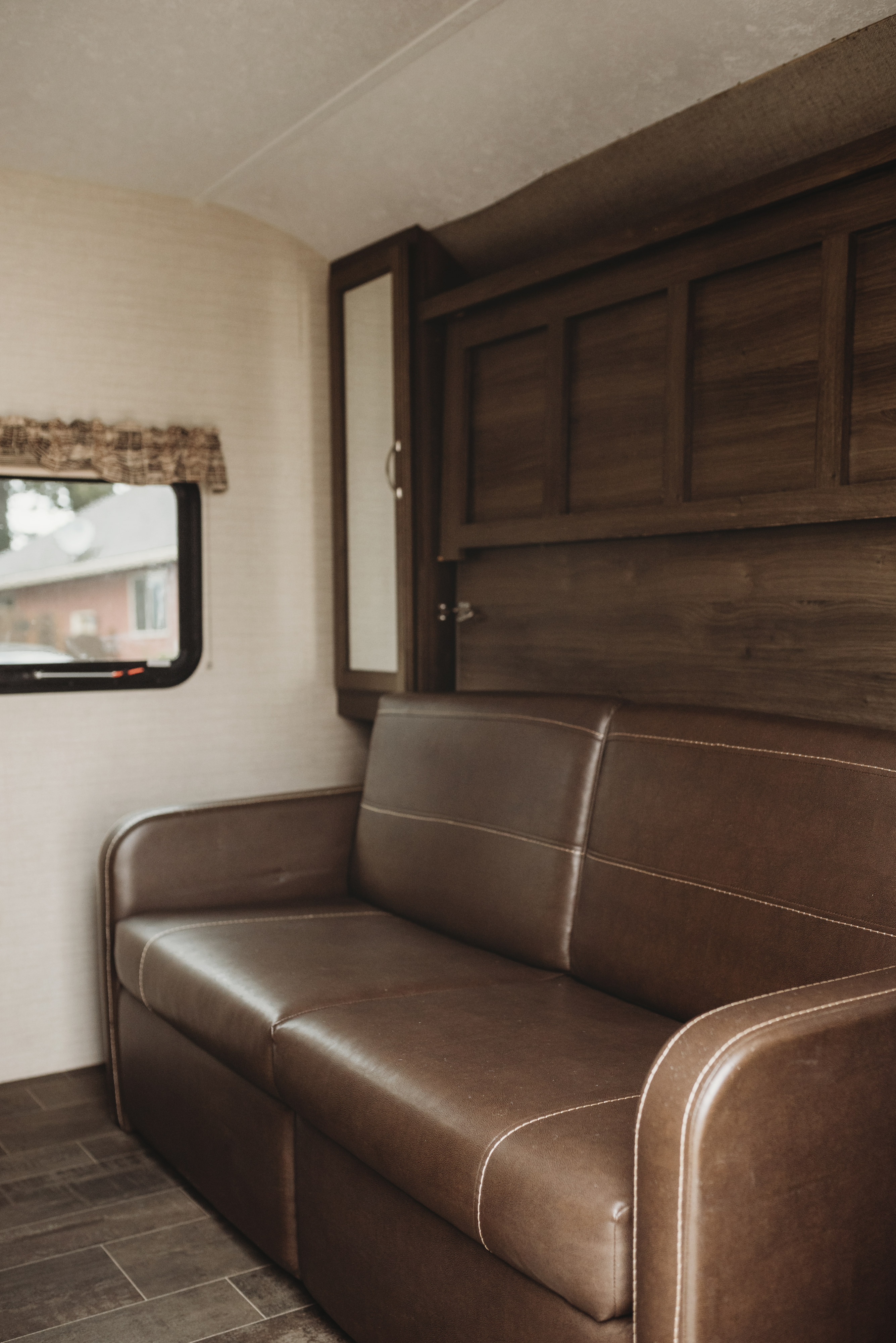 Cozy RV rental interior featuring a brown leather sofa with white stitching, dark wood cabinets, and a compact window.. Keystone RV Bullet Ultra Lite 2019