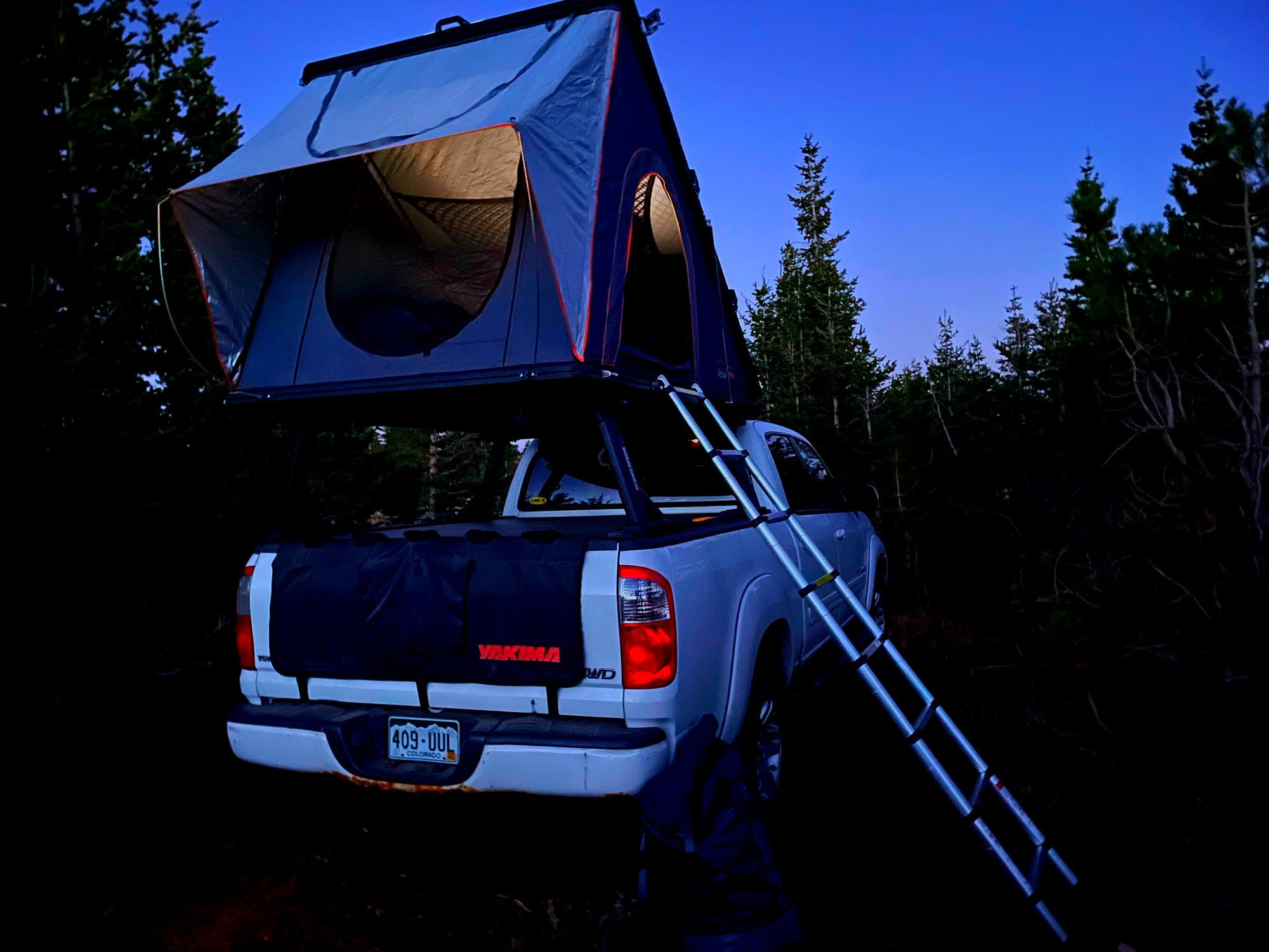A white pickup truck with a lit rooftop tent and ladder, an excellent RV rental option for overlanding adventures at dusk.. American Dream Toyota 2006