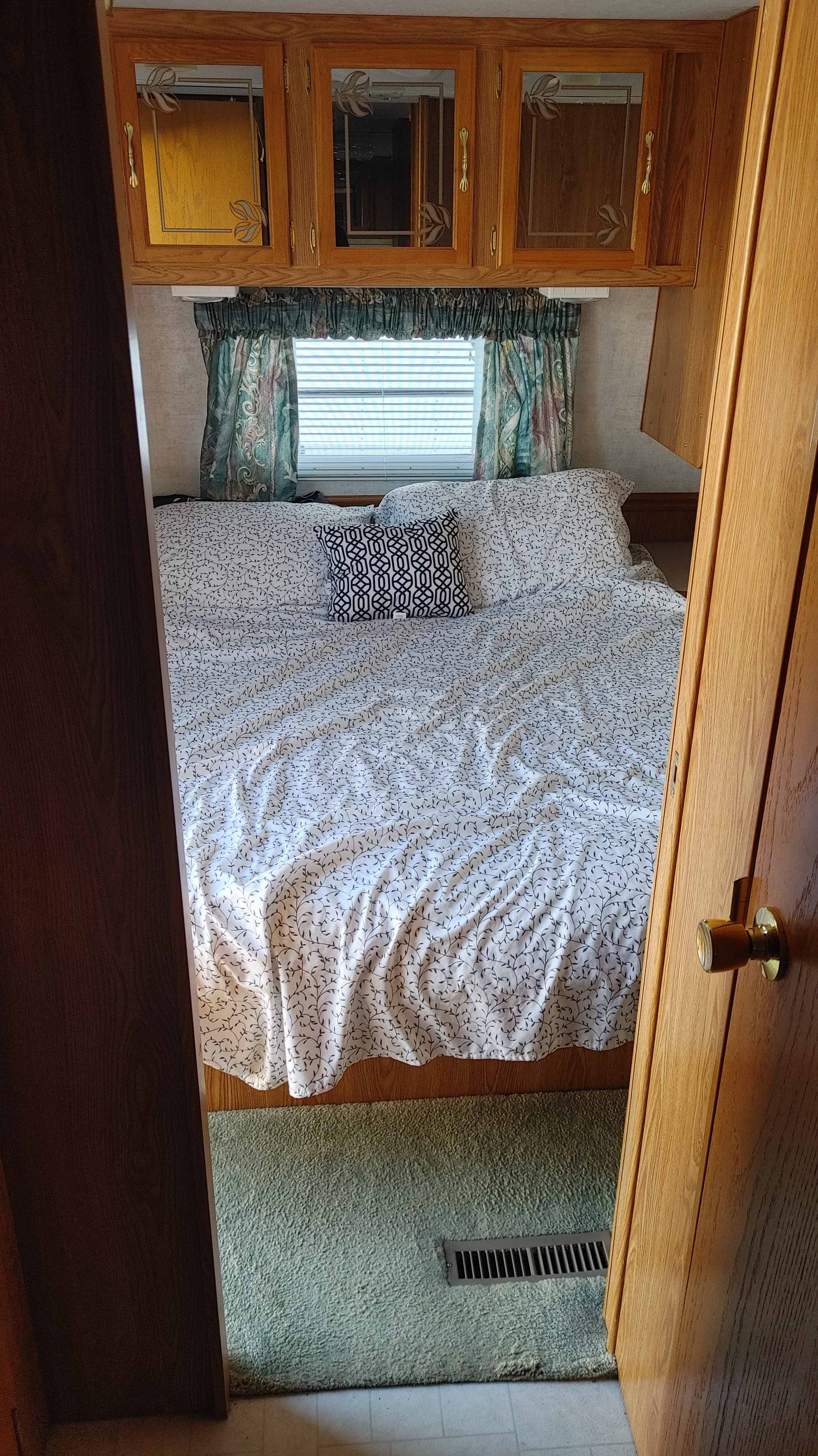 Inside an RV rental: a compact bedroom featuring a bed with patterned sheets, overhead cabinets, and a window with blinds.. Sunnybrook Sunnybrook Trailer 2000