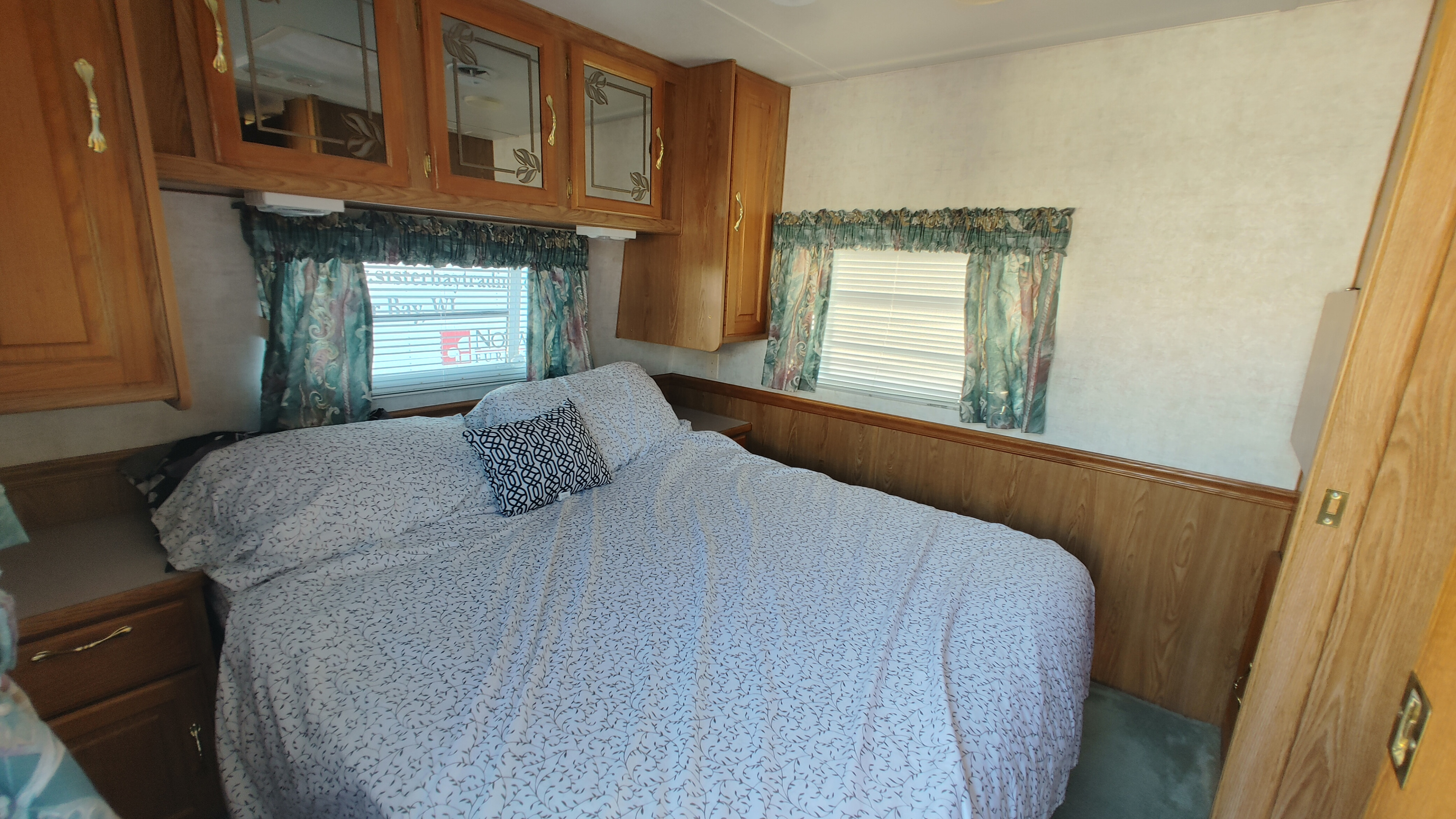 Cozy RV rental bedroom with a comfortable bed, overhead storage cabinets, and a window with patterned curtains.. Sunnybrook Sunnybrook Trailer 2000