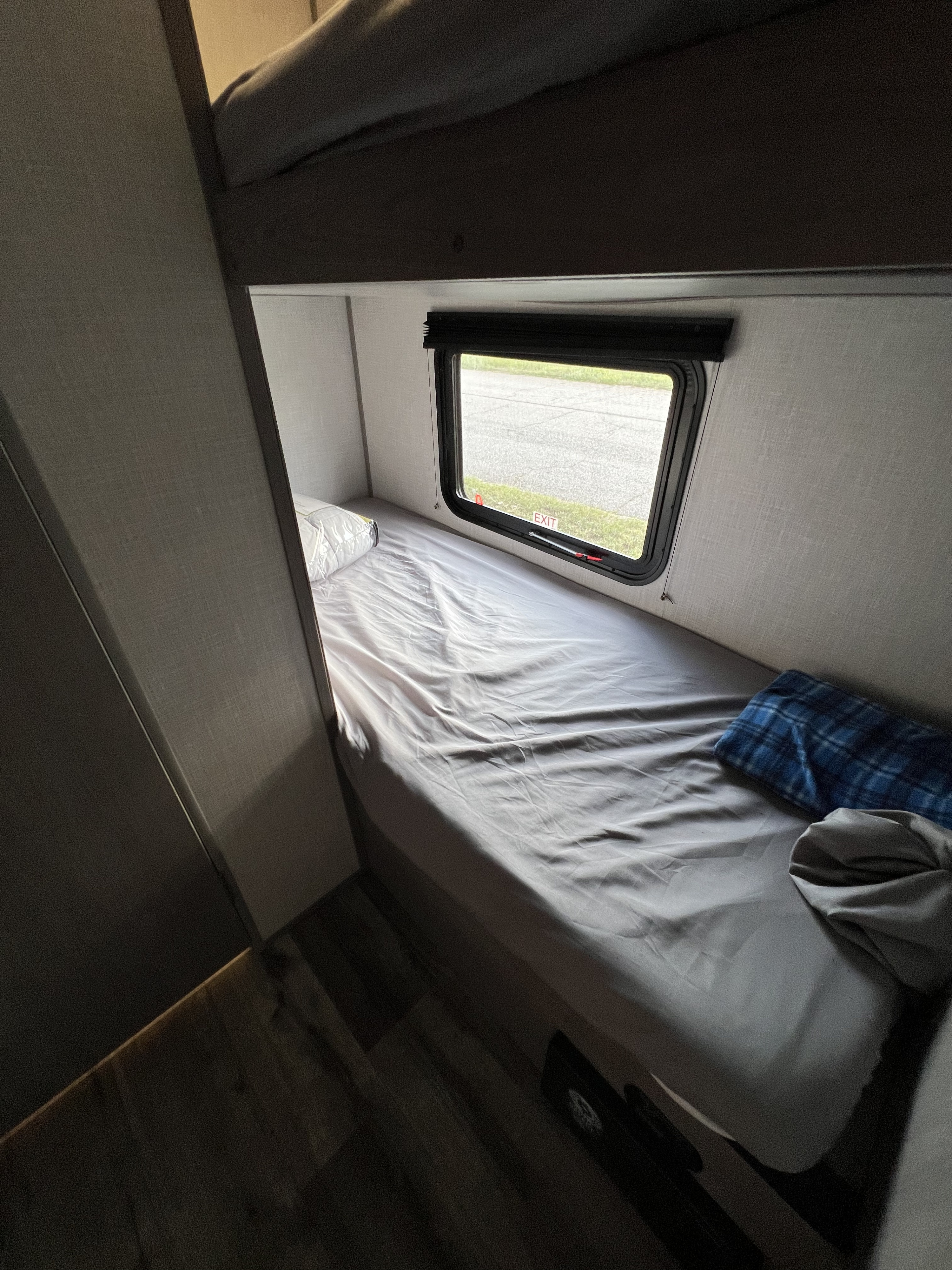 This RV rental features a comfortable bunk bed setup. The lower bunk has grey sheets, a pillow, and a window with an "EXIT" sticker.. Dutchmen Coleman Lantern LT 2022