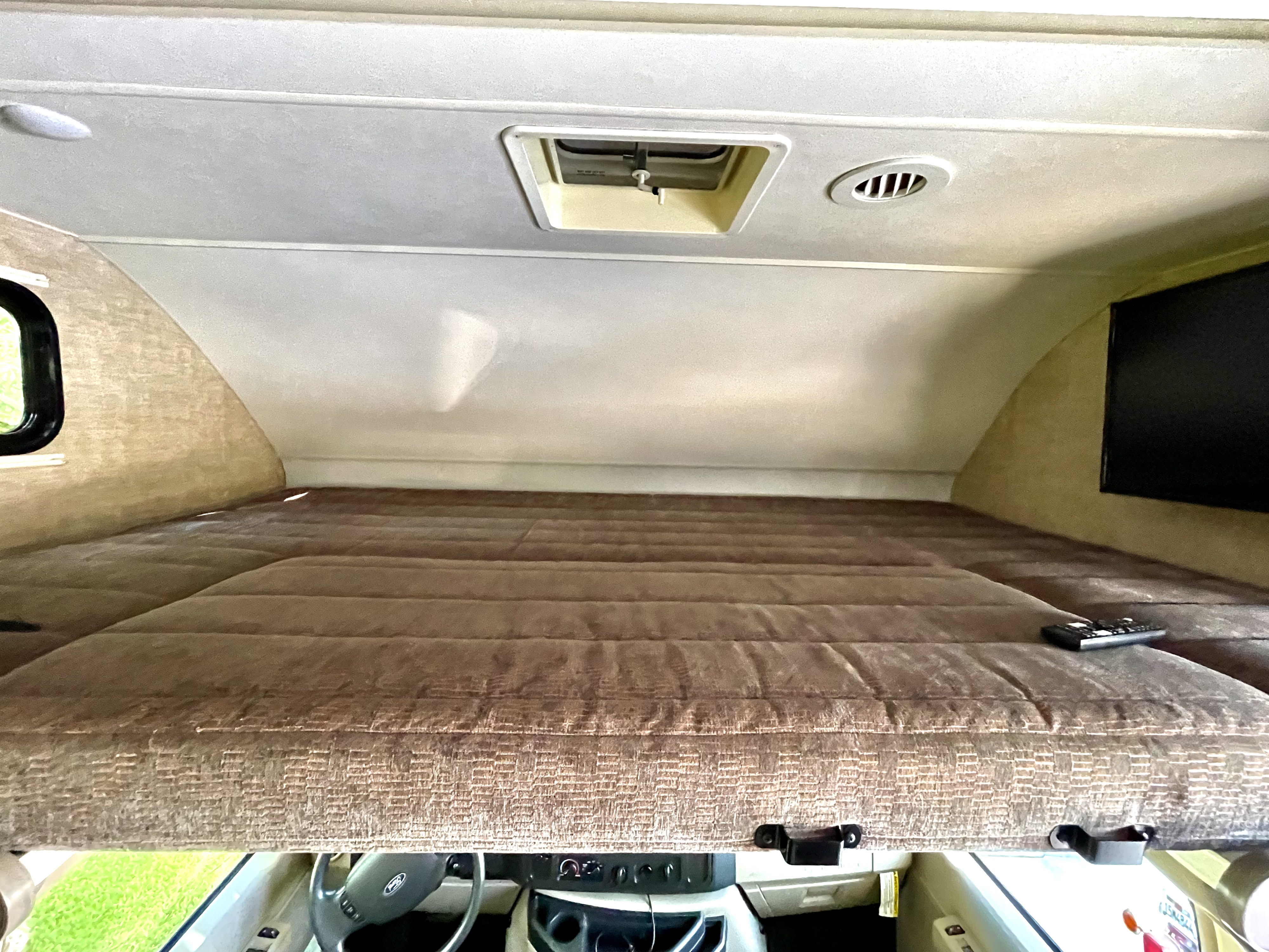 RV rental's cab-over bunk features a quilted brown mattress, overhead vent, skylight, and TV. Driver's cockpit below.. Winnebago Minnie Winnie 2019
