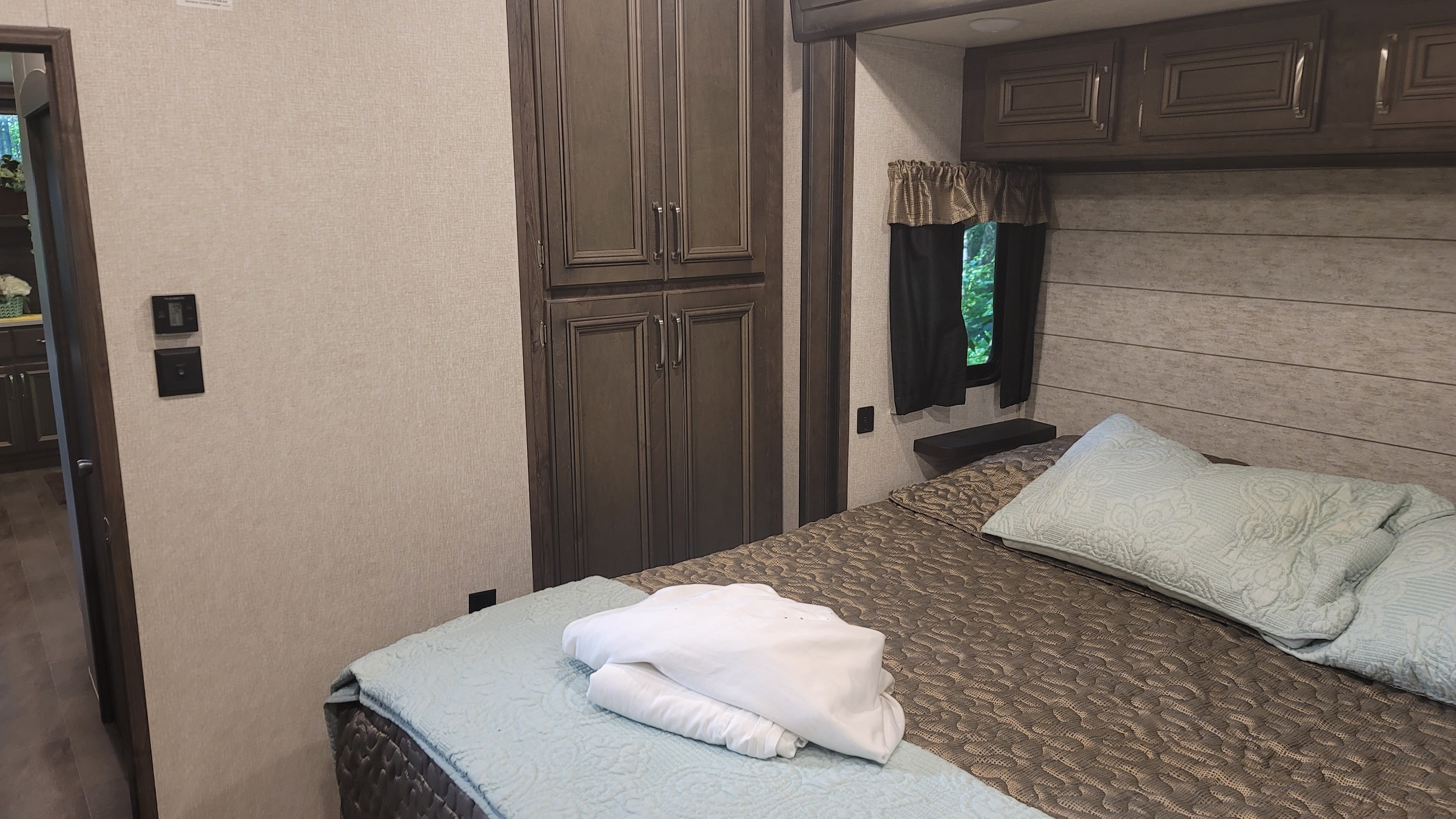 Cozy RV rental bedroom featuring dark wood cabinets, a comfortable bed with light bedding, and a window with green views.. Keystone RV Montana High Country 2019