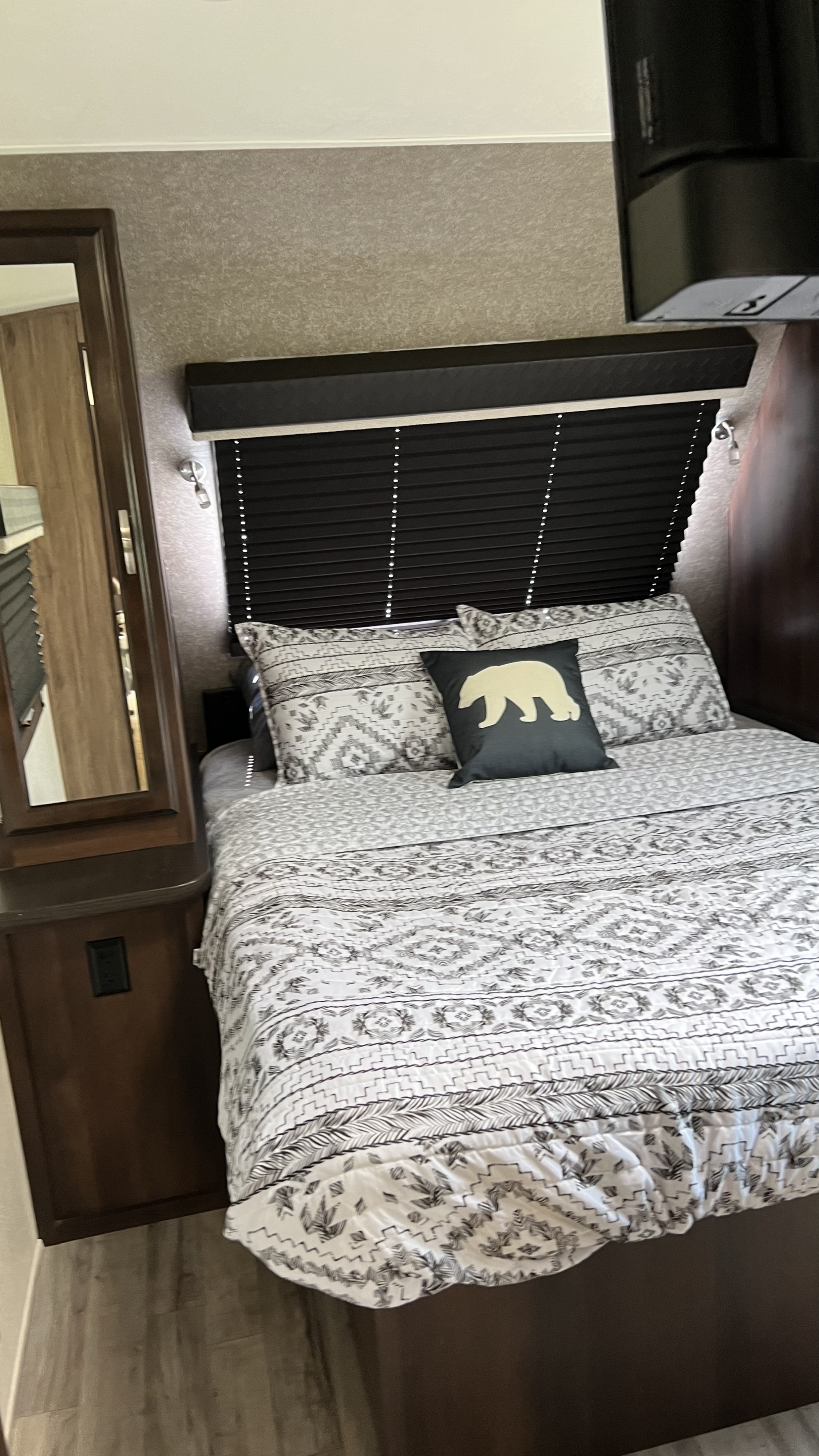 This RV rental bedroom features a neatly made bed with white and gray patterned bedding, a bear pillow, and a built-in vanity.. Jayco White Hawk 2019