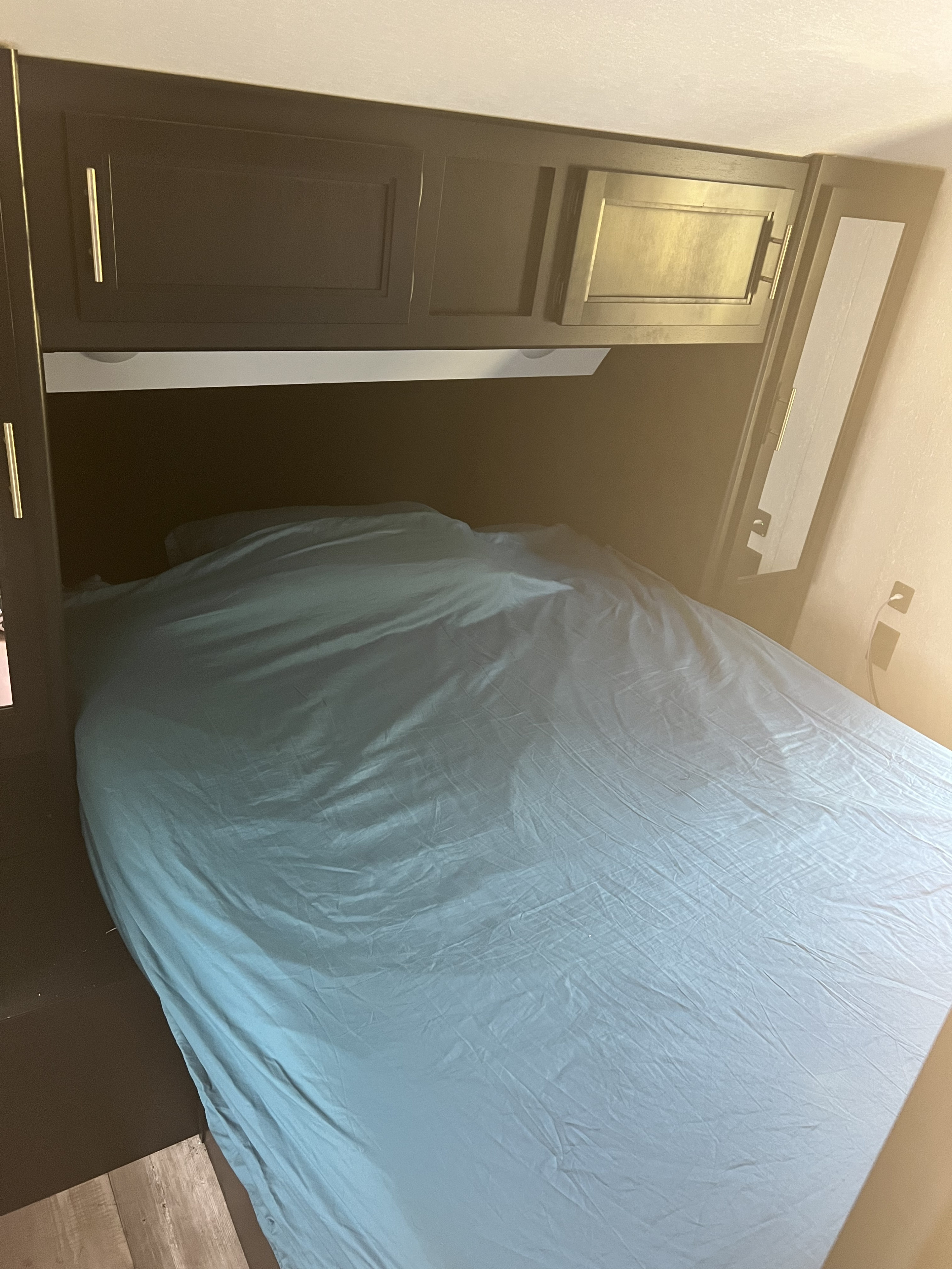 Inside an RV rental: a compact bedroom with a bed, teal sheet, and dark overhead cabinets.. Forest River Cherokee 2021
