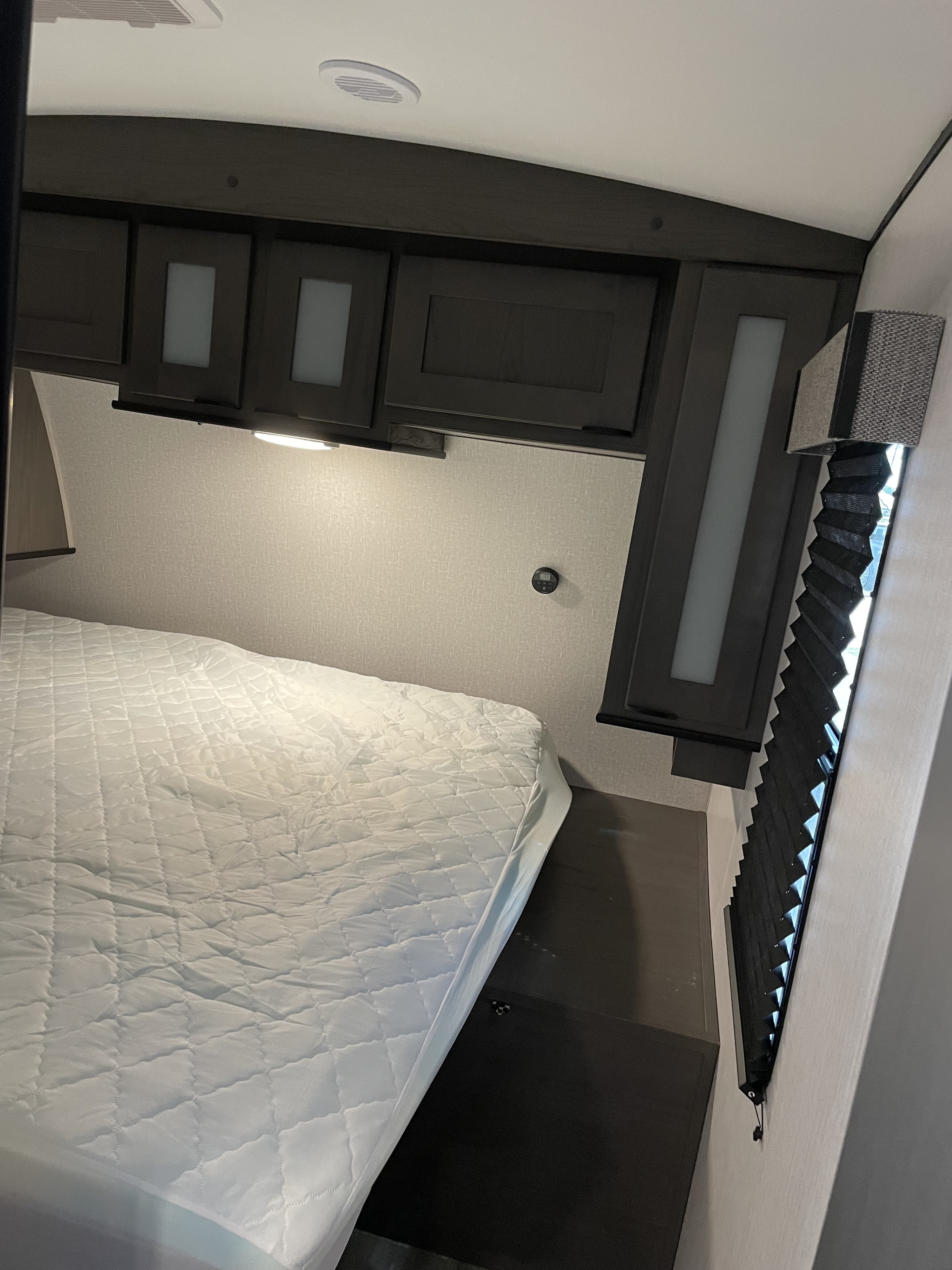 Cozy RV rental bedroom featuring a white quilted bed, dark overhead storage with integrated lighting, and pleated window blinds.. Heartland Mpg 2022